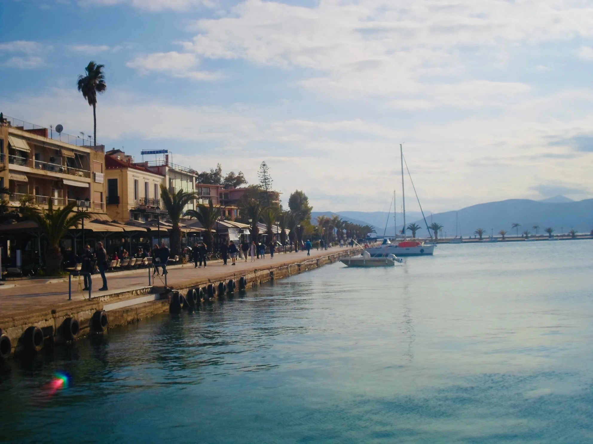 A seaside promenade with people walking along the waterfront, palm trees, colorful buildings with cafes and shops, and boats docked in calm water under a partly cloudy sky.