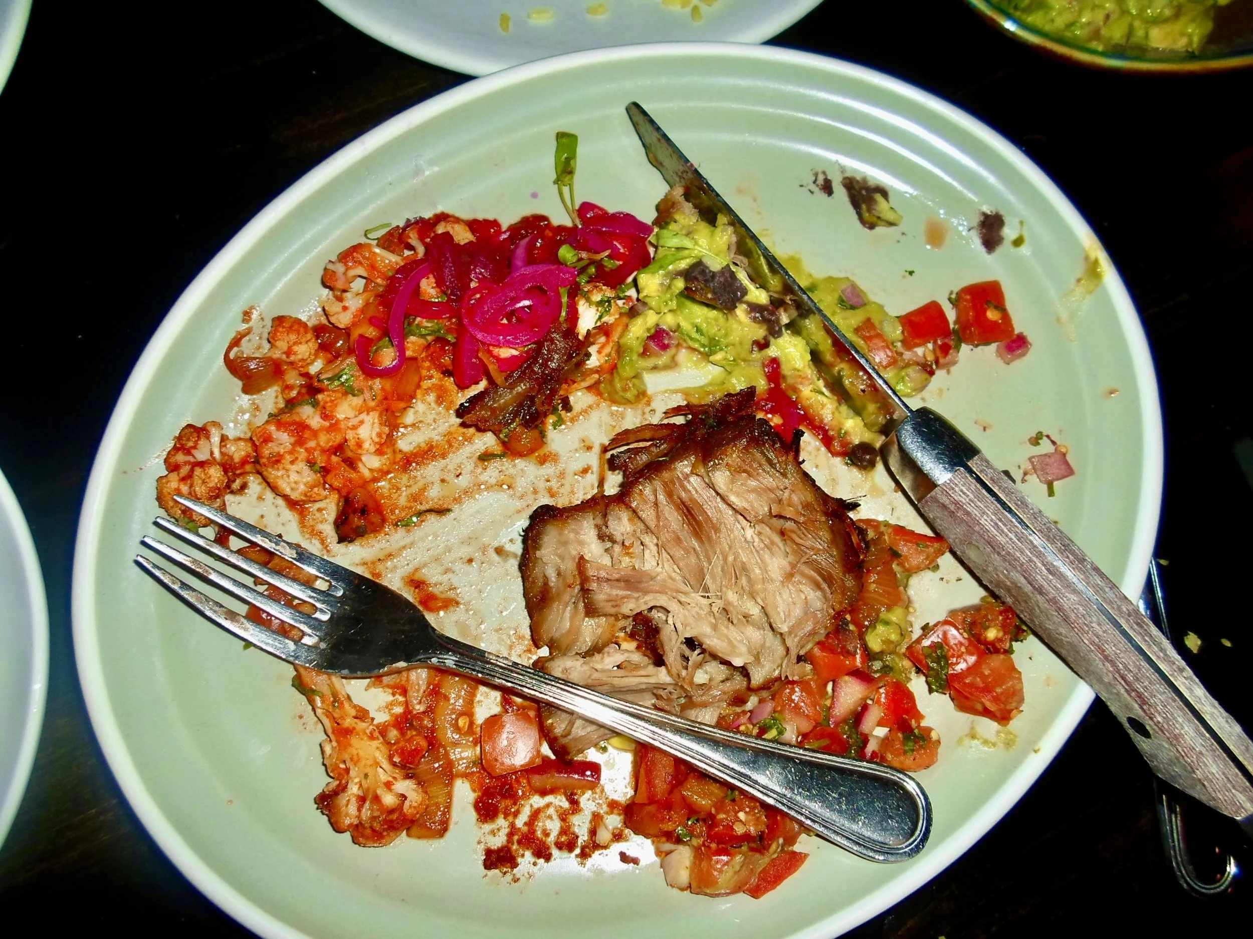 Empty plate with leftover food, including a piece of cooked meat, chopped vegetables, and scattered bits of food, with a fork and knife resting on it.