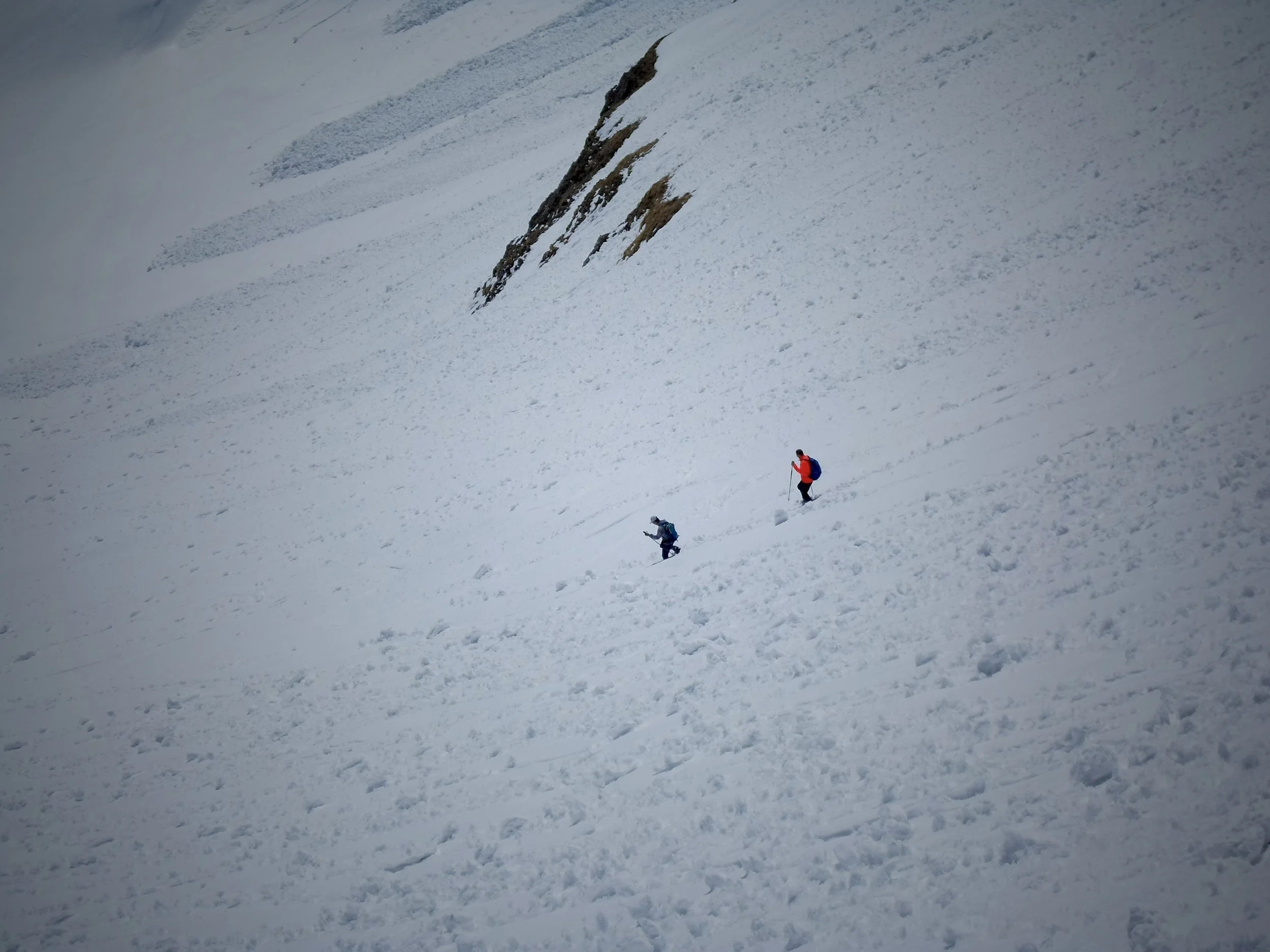 Two hikers trekking through snow on a steep, snowy mountain slope, with rocky outcrops in the background.