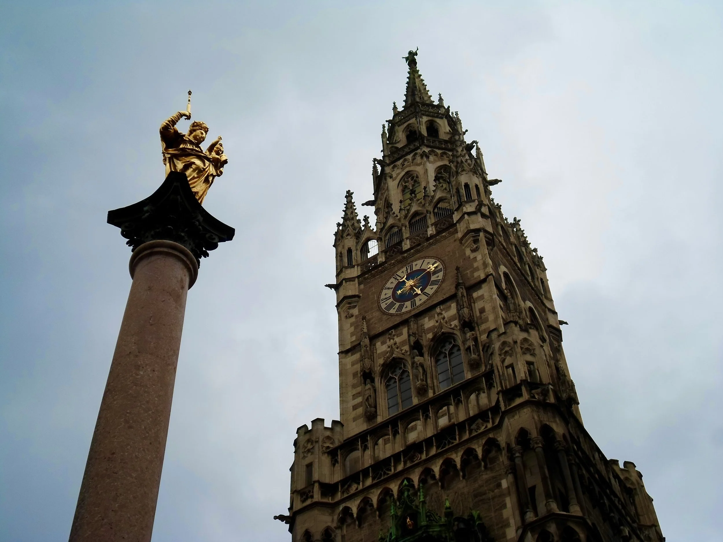 A gothic-style clock tower and a golden statue of a figure on a tall column against a cloudy sky.