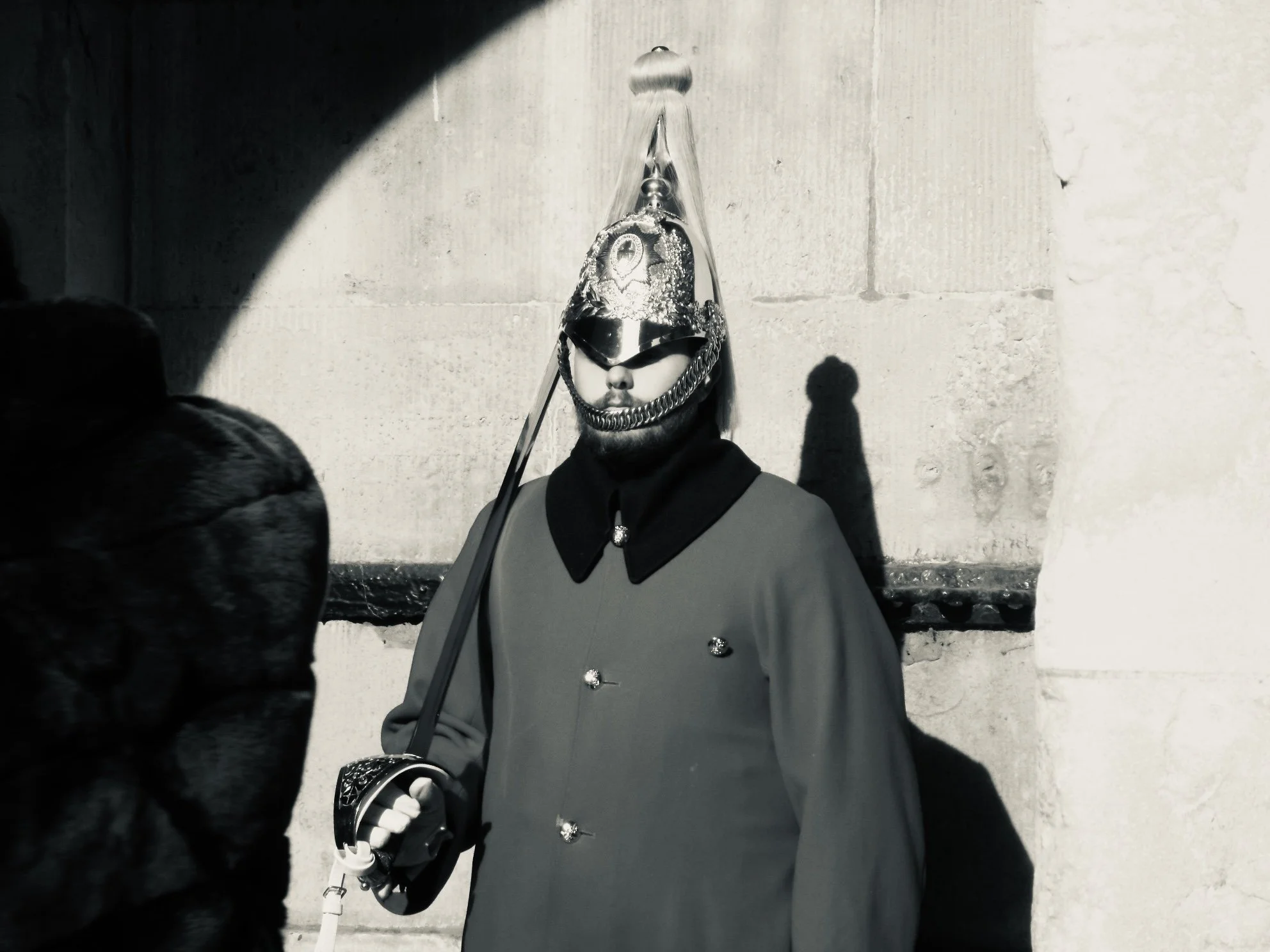 A man in a military-style uniform wearing a shiny helmet and sunglasses standing against a concrete wall, with a shadow cast on the wall.