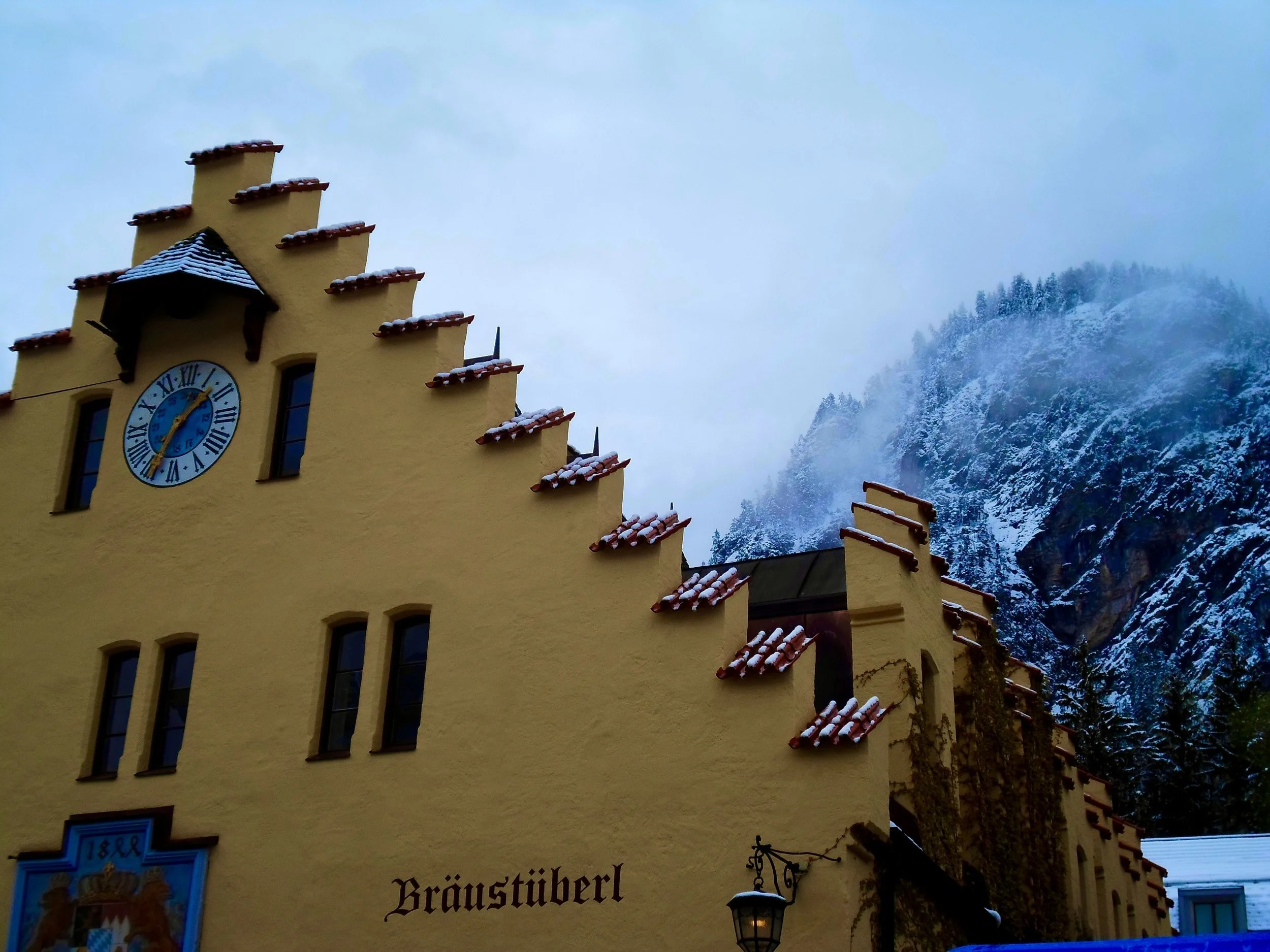 A yellow building with a clock tower, red-tiled roof and snow-covered mountains in the background.