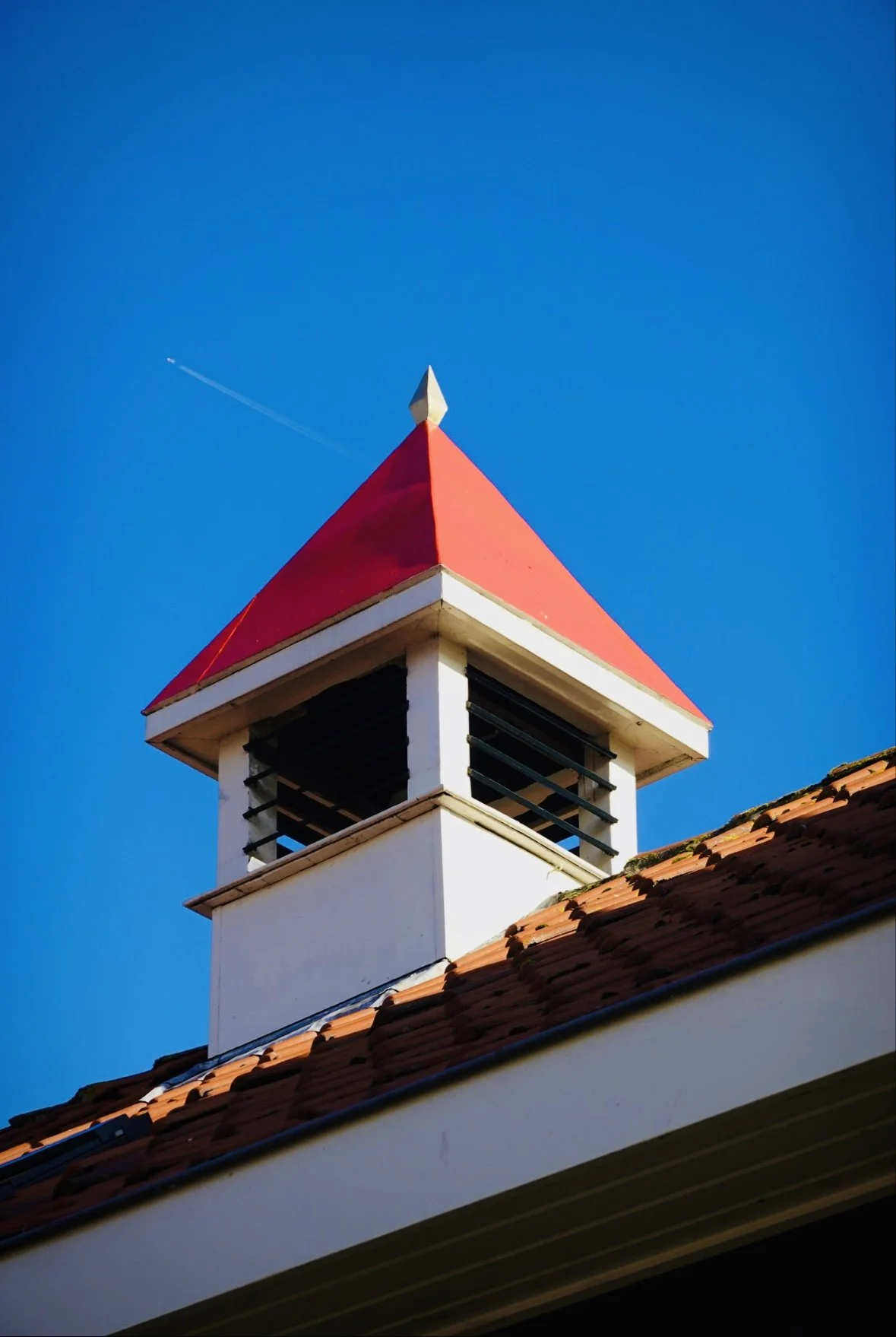 Close-up of a building's cupola with a red, pyramid-shaped roof and a white structure below, set against a clear blue sky with a small contrail.