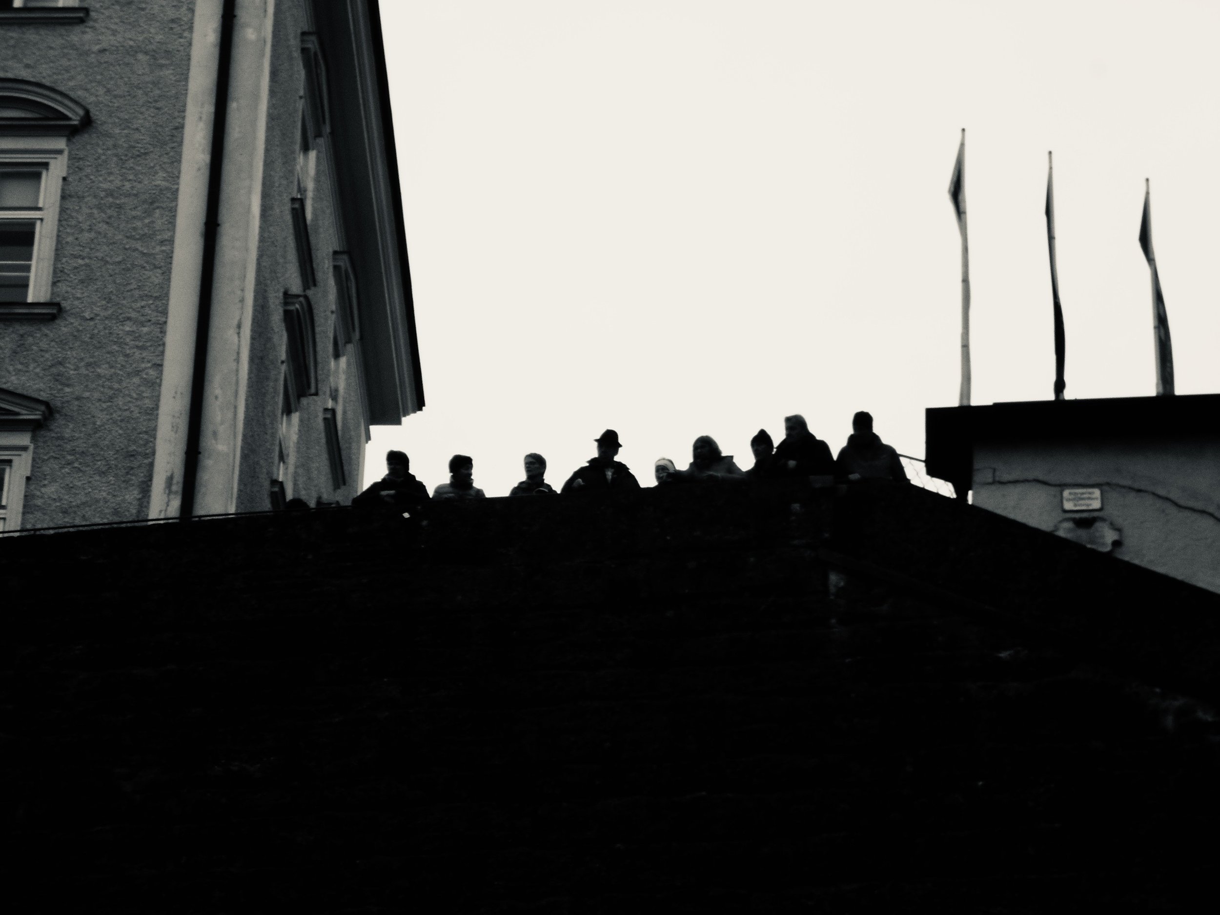 Silhouettes of people sitting on a rooftop against a light sky, with a building on the left and flags on the right.