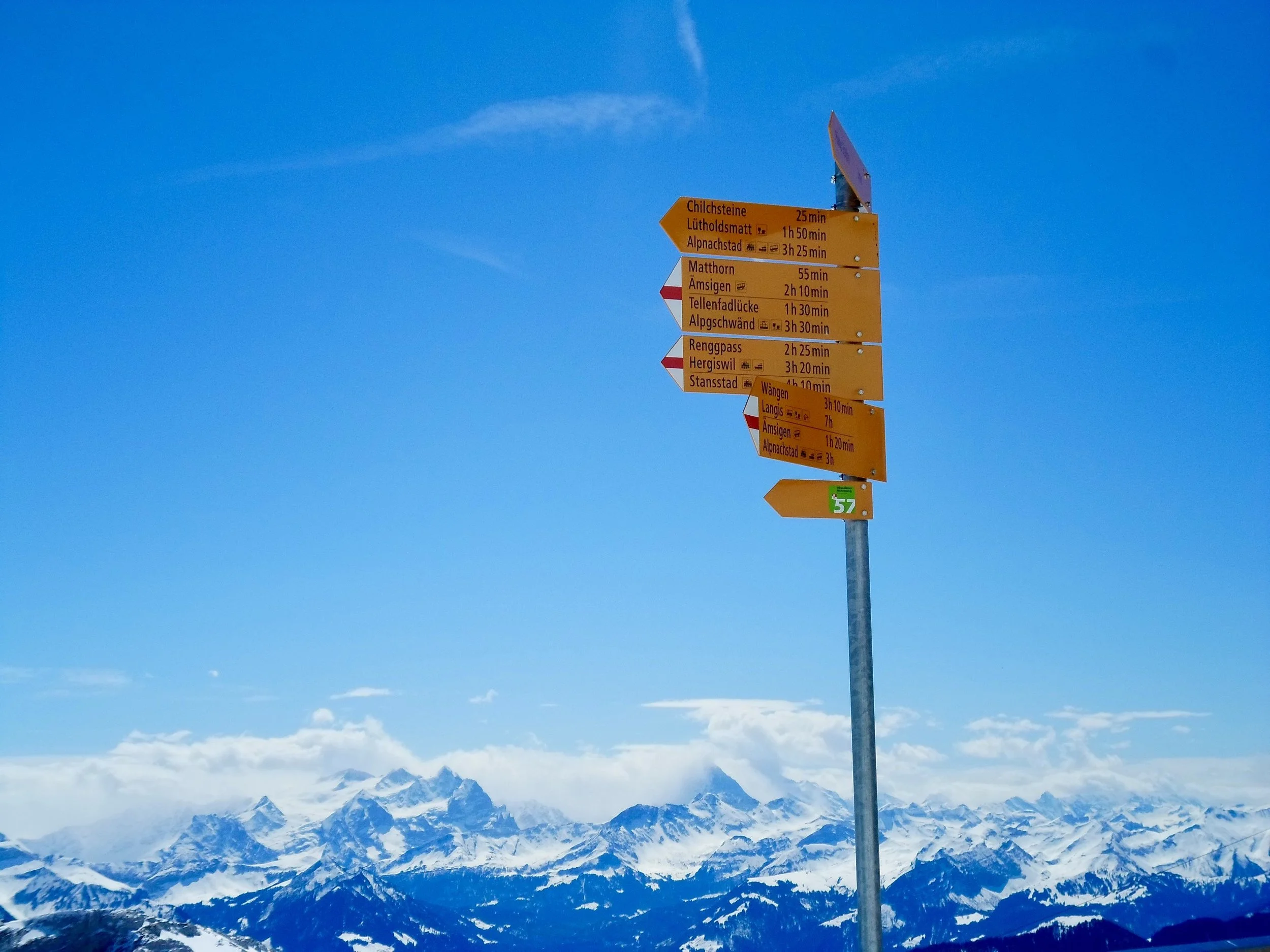 Mountain ski resort with a directional signpost against snowy mountains and a clear blue sky.