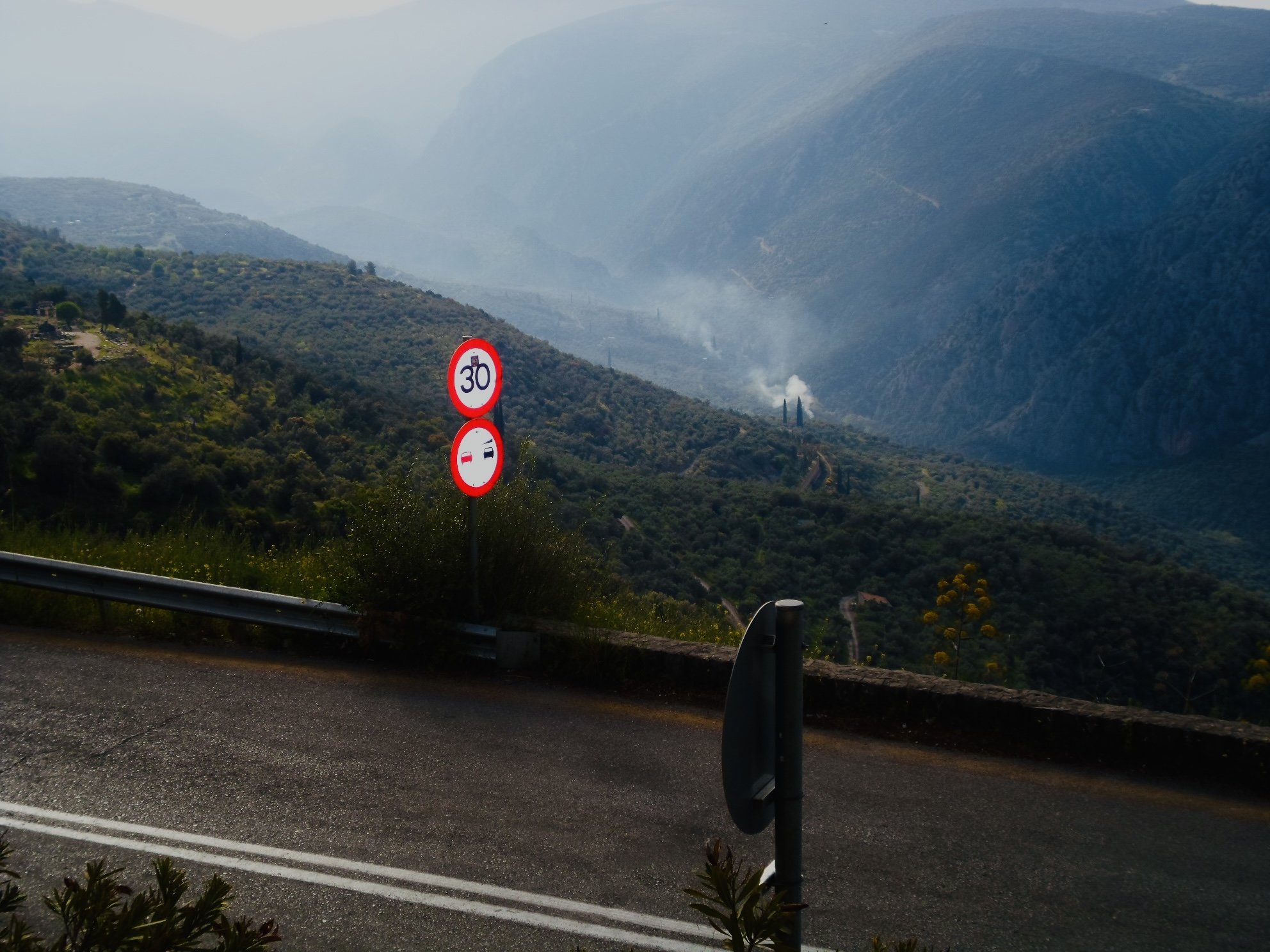 Mountain road with traffic signs indicating a speed limit of 30 and no overtaking, overlooking a valley with distant smoke or fog rising from the hills.