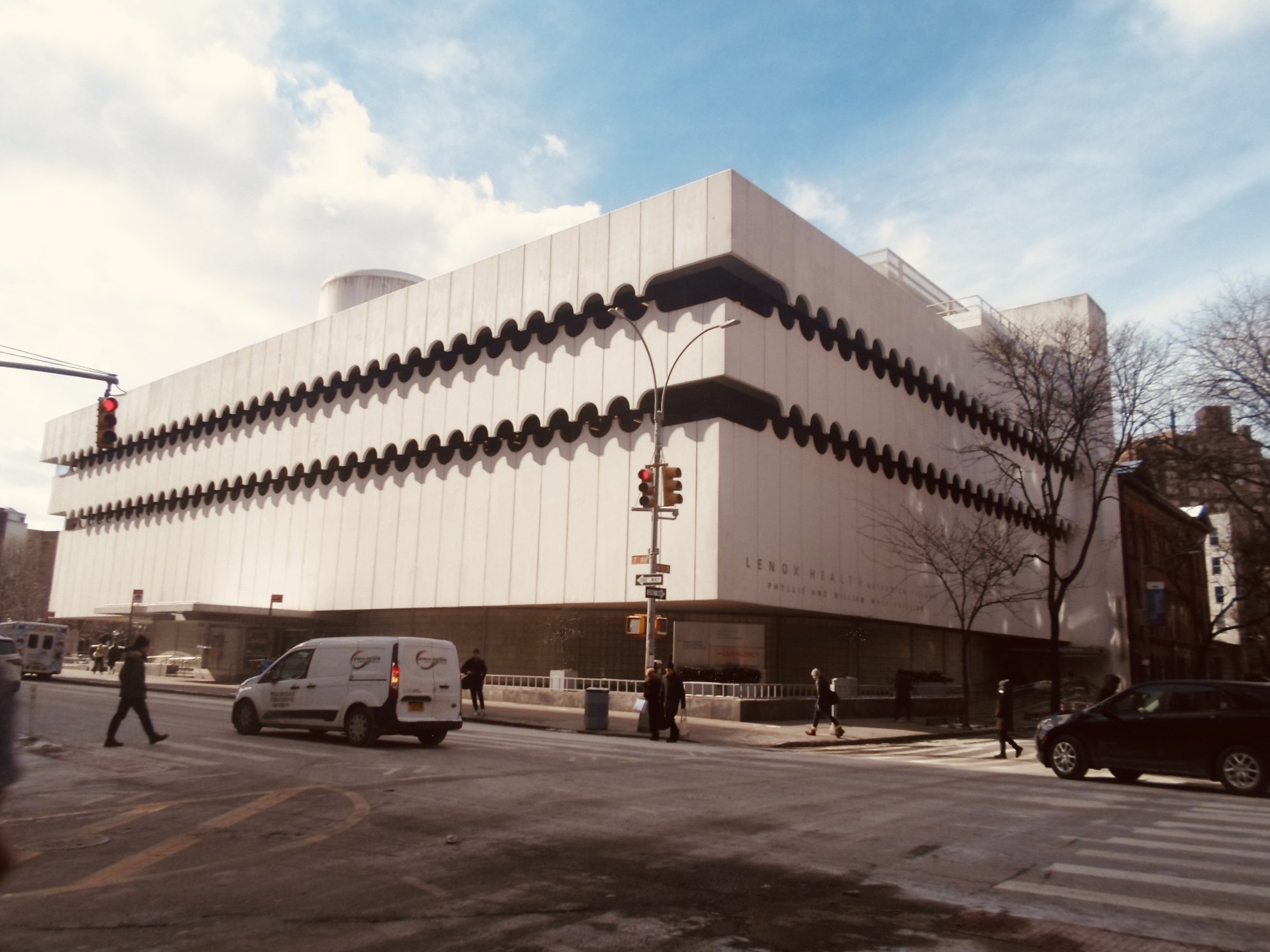 The Lenox Health building, a modern white structure with black scalloped accents along the top and sides, is seen on a city street corner with pedestrians crossing and cars passing by. Bare trees are in front of the building, and the sky is partly cl