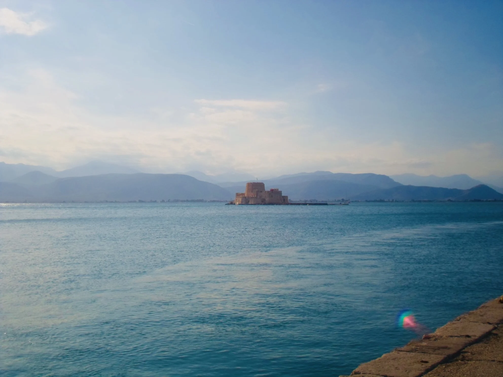 A body of water with a historic fortress or castle on a small island in the distance, mountains in the background, and a partly cloudy sky.