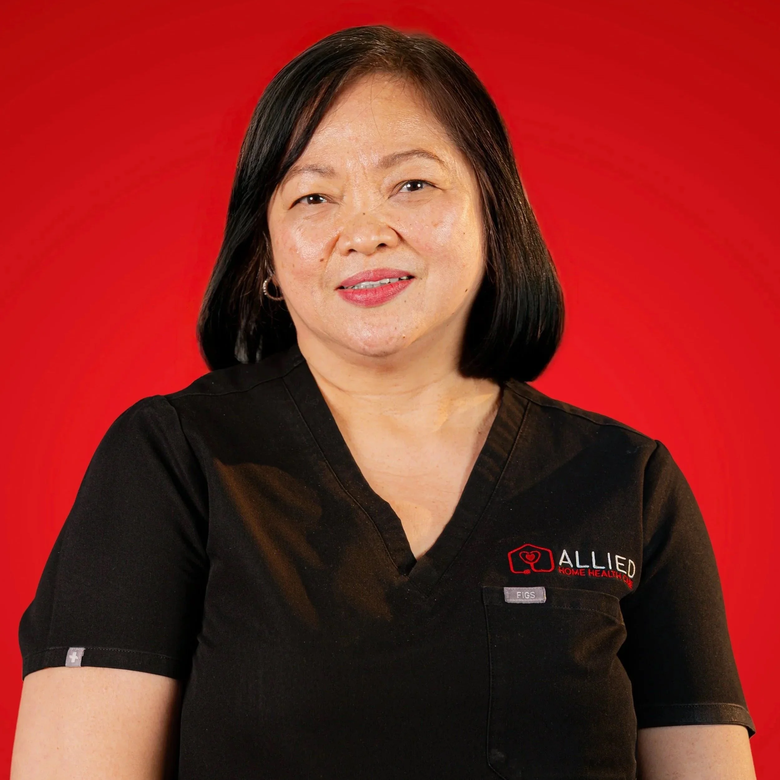 A woman with black hair wearing a black uniform with 'ALLIED HOME HEALTH' logo, standing against a red background.