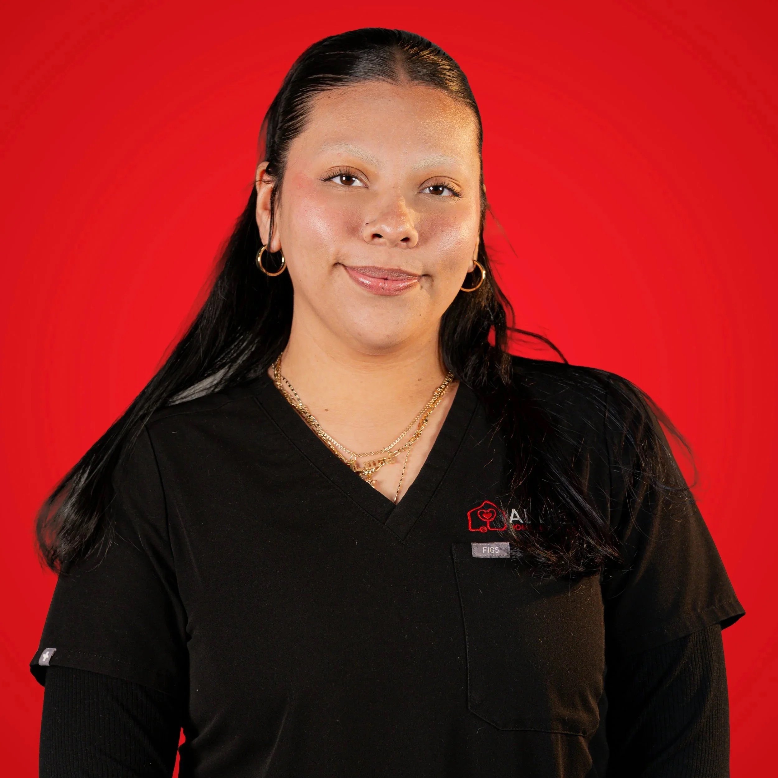 A woman with long black hair, wearing gold jewelry, and a black uniform with a red and white logo, standing against a red background.