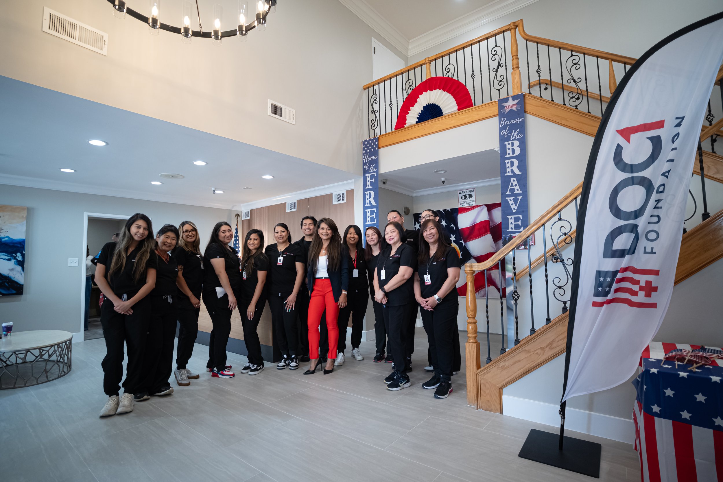 Group of people standing inside a building decorated for a patriotic event, with American flags, banners reading 'Home of the Free Because of the Brave,' and a large red, white, and blue 'FOOD! FOUNDATION' flag on the right.