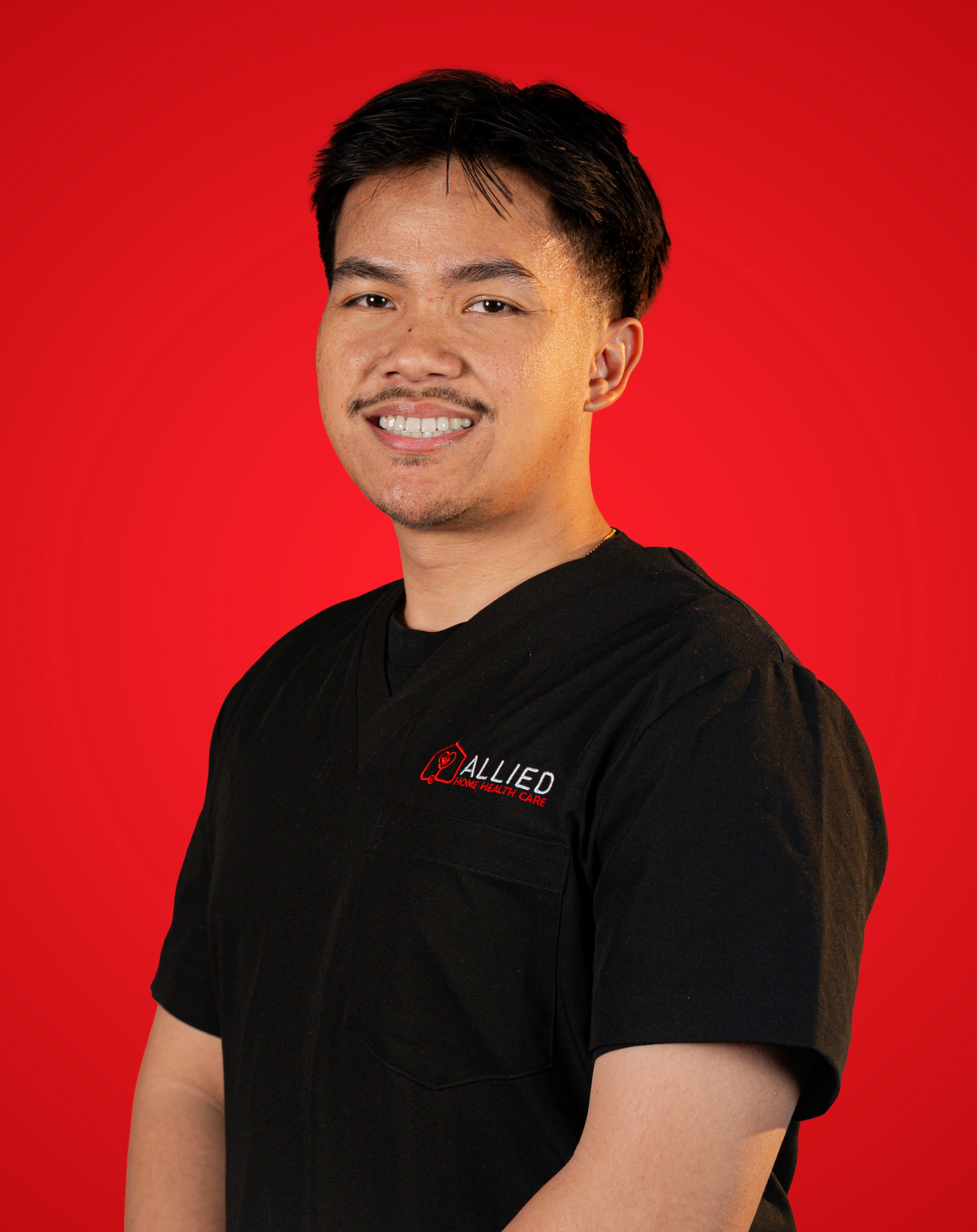 A man smiling, wearing a black medical scrub top with a logo and text that read 'ALLIED HOME HEALTH CARE', standing against a red background.