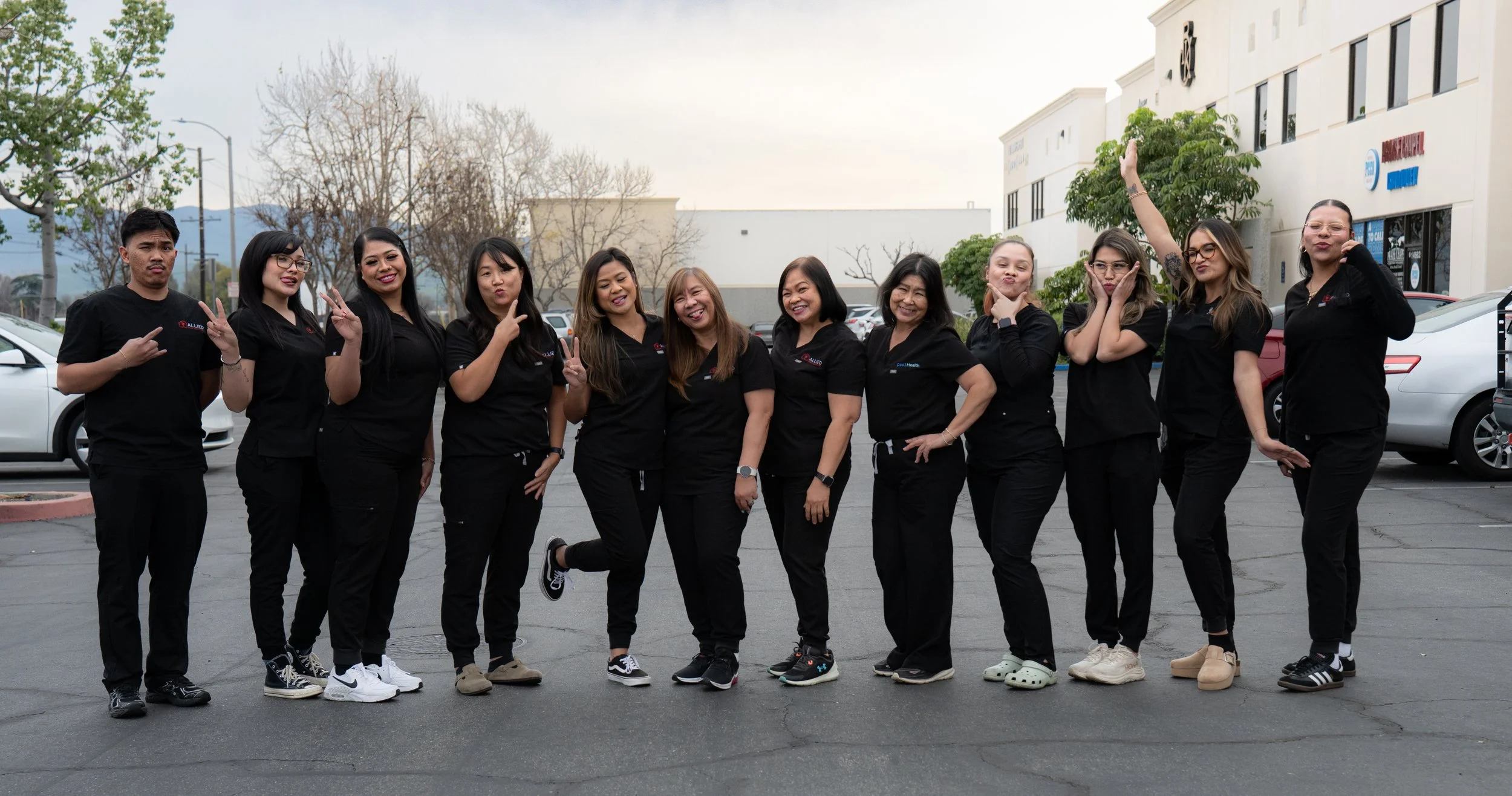 Group of nine women and one man standing outdoors in a parking lot, all wearing black uniforms, some making peace signs, smiling, and striking playful poses.