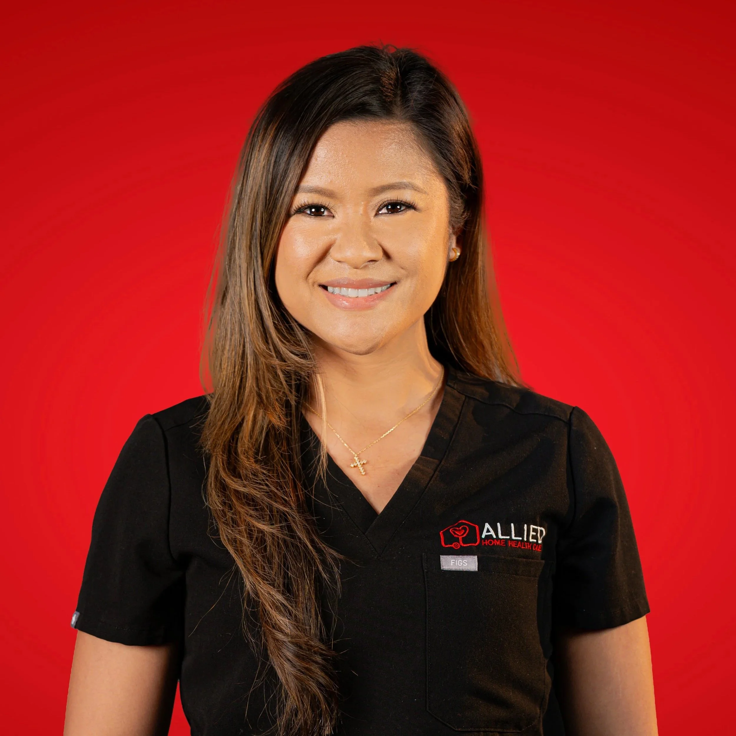 A woman with long, brown hair smiling, wearing a black medical uniform with 'ALLIED Home Health Care' logo on the left chest, standing against a red background.