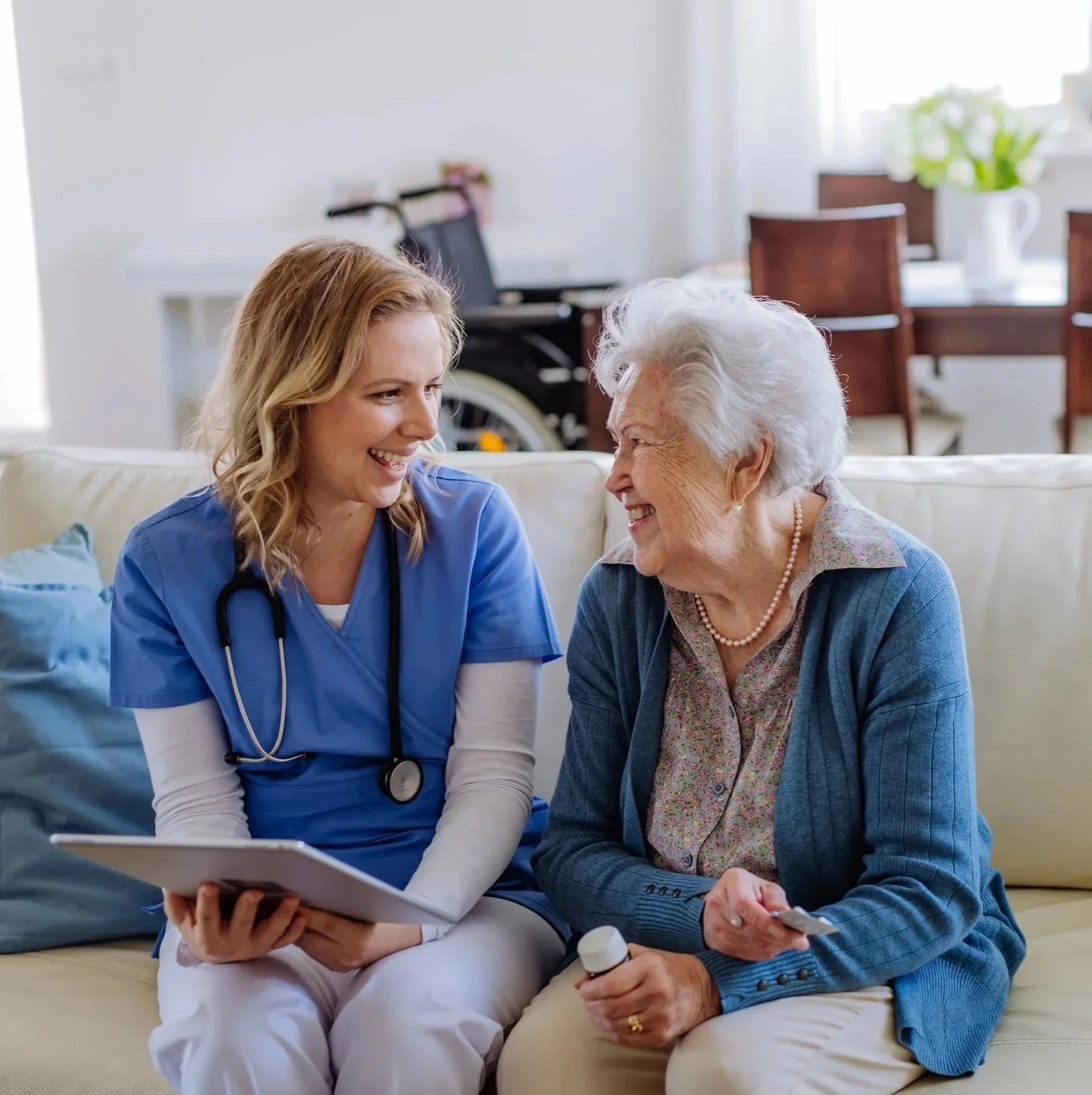 Nurse in blue scrubs smiling and talking with elderly woman sitting on a sofa, holding medicine bottle, in a bright living room.