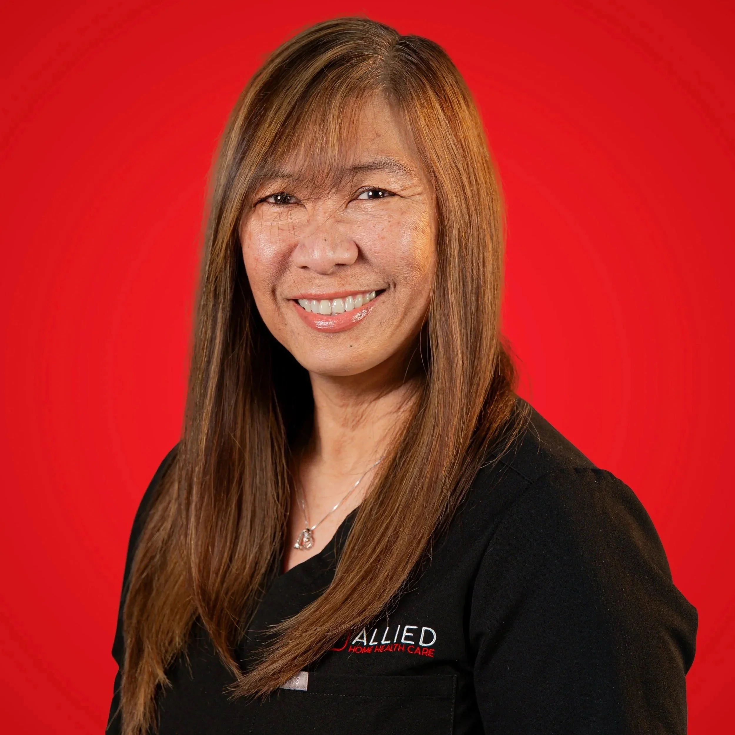 Portrait of a smiling woman with long brown hair, wearing a black top with a logo on it, against a red background.