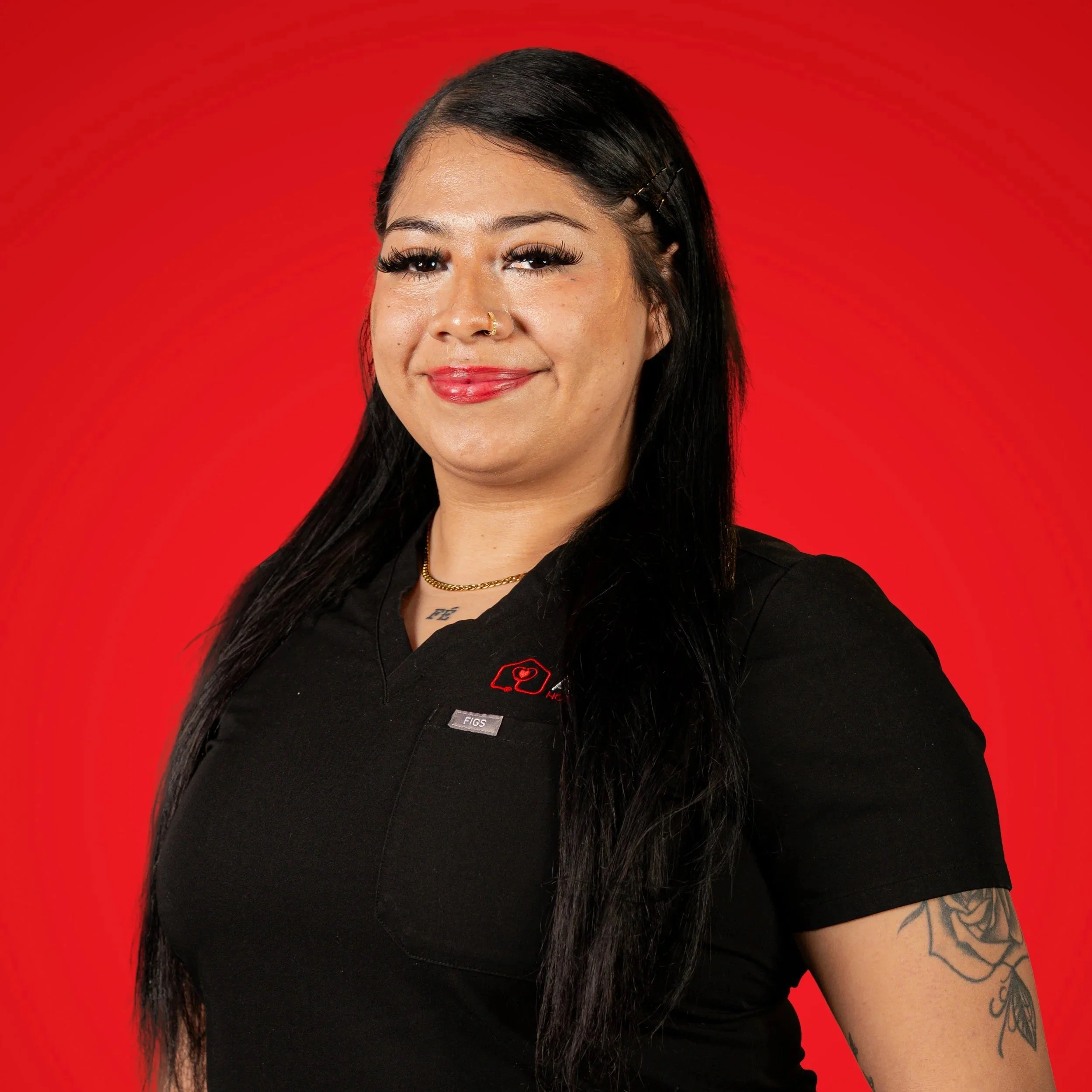 A woman with long black hair, wearing a black shirt with red logo, standing against red background, smiling at the camera.