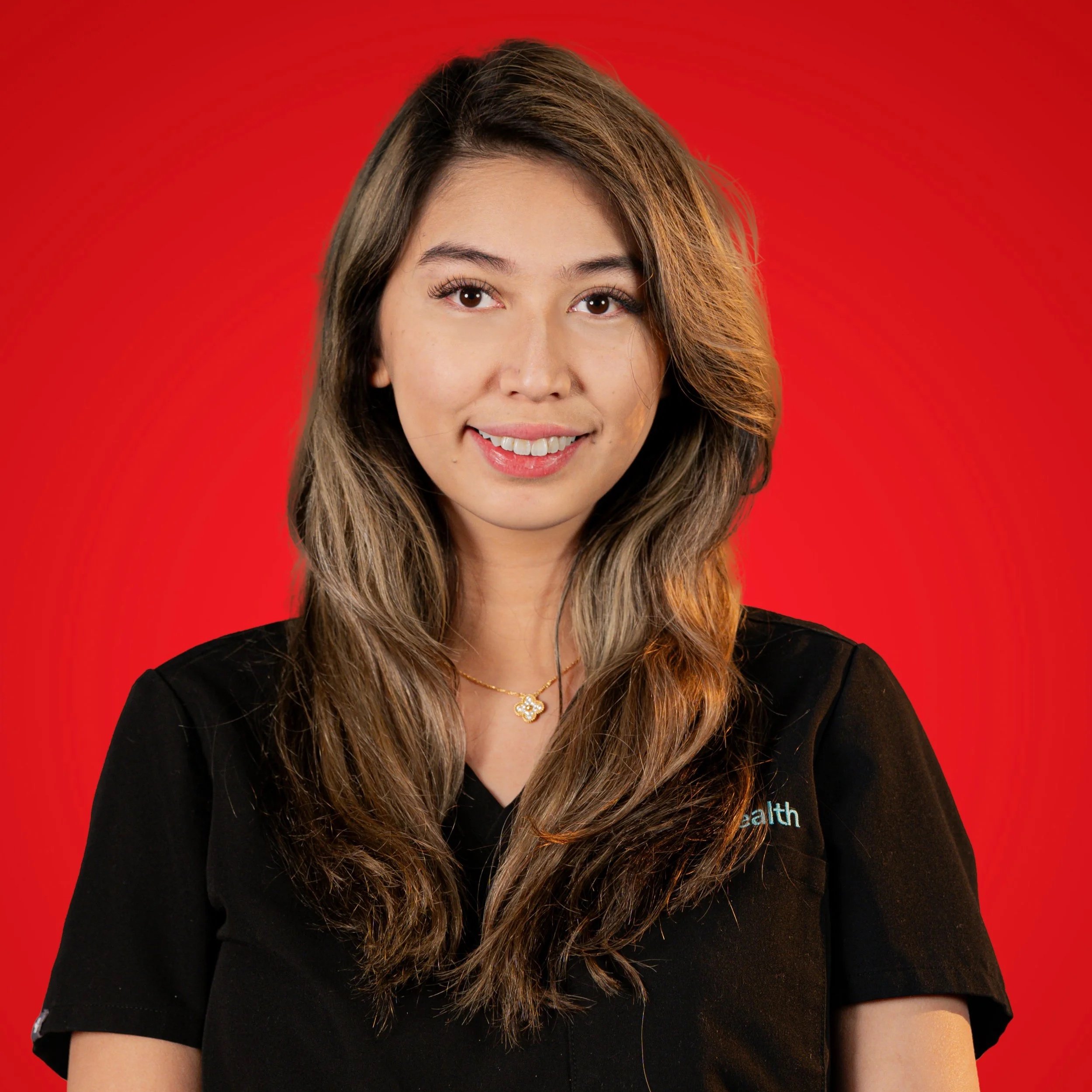 Portrait of a young woman with long wavy brown hair, wearing a black top and a gold necklace, smiling against a red background.