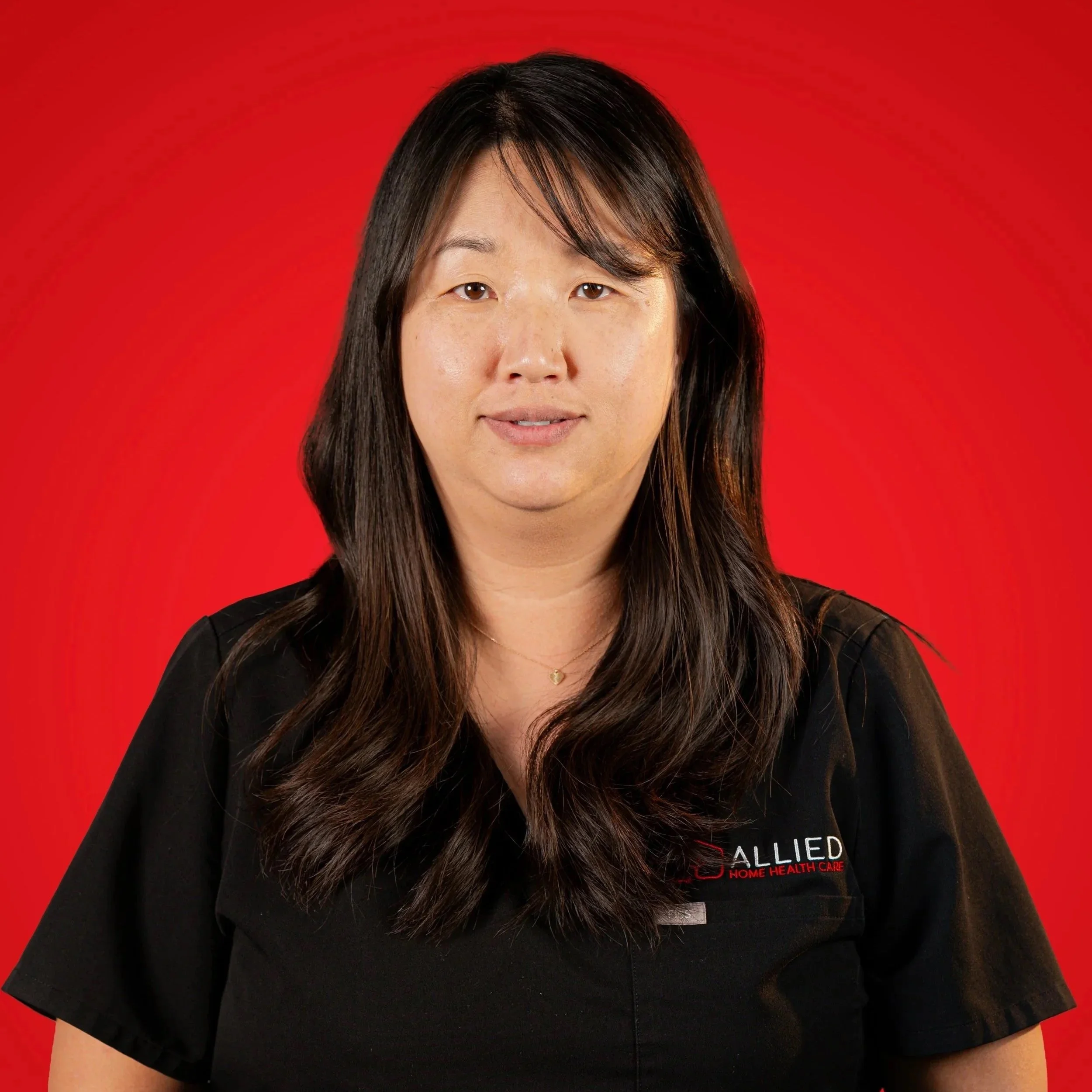 Portrait of a woman with long dark hair, wearing a black shirt with 'ALLIED HOME HEALTH CARE' logo, against a red background.