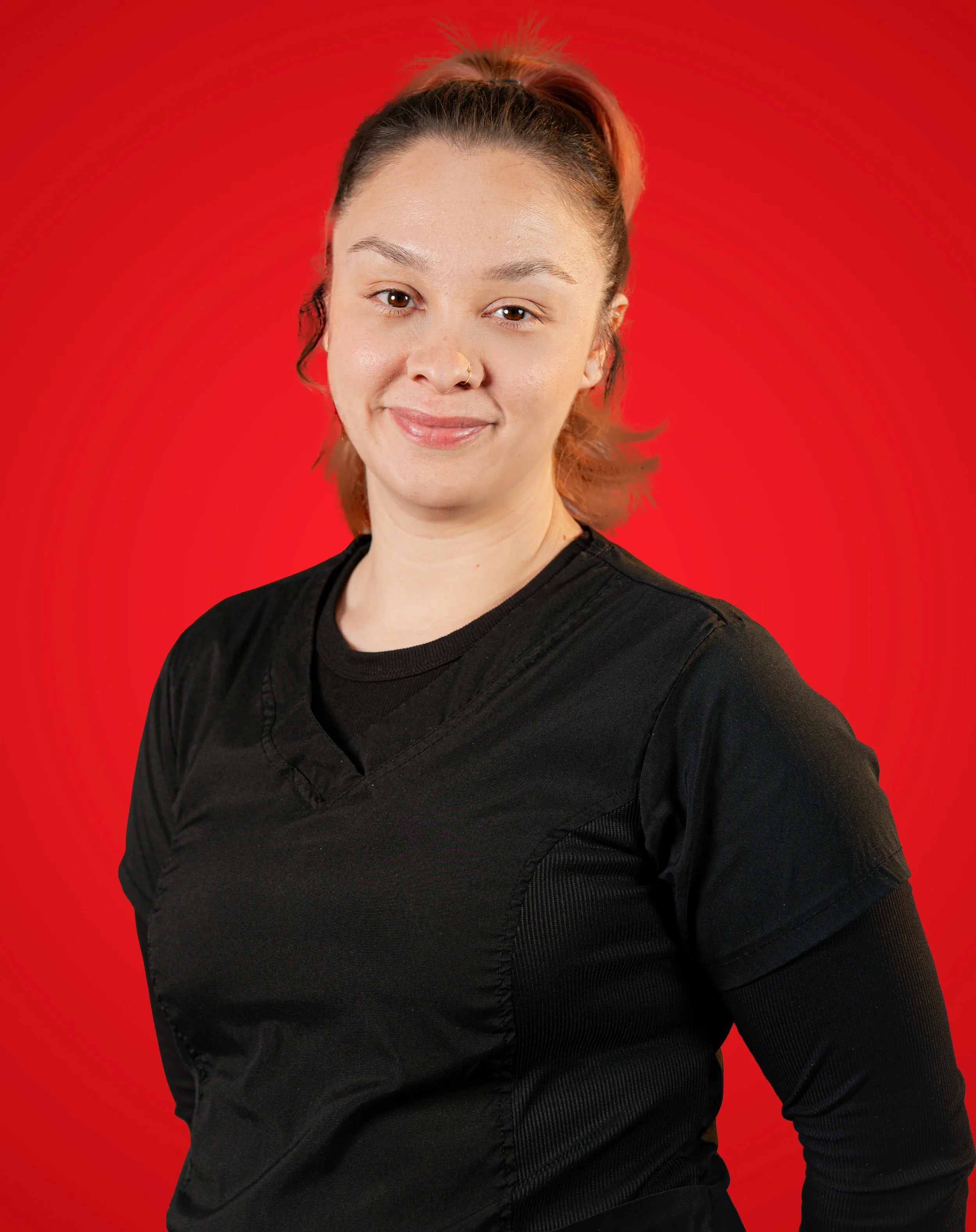 Portrait of a woman with light skin, red hair tied back, wearing black medical scrubs, standing against a red background.