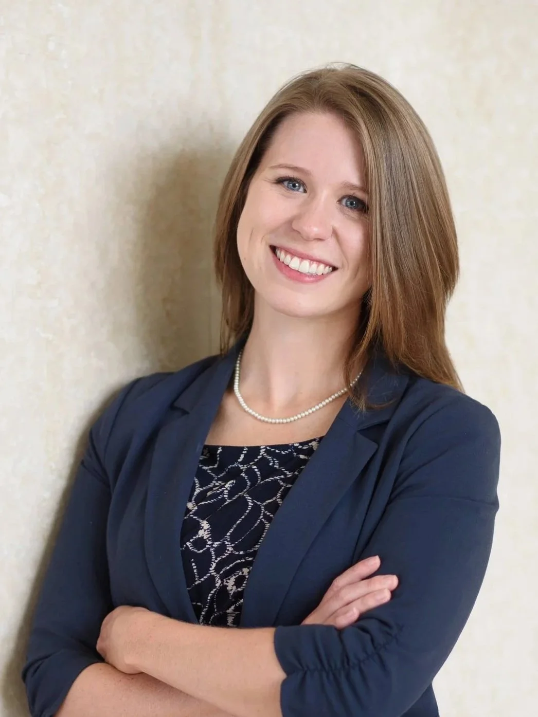 A smiling woman with shoulder-length reddish-brown hair, wearing a navy blazer, black patterned top, and pearl necklace, standing against a neutral background.