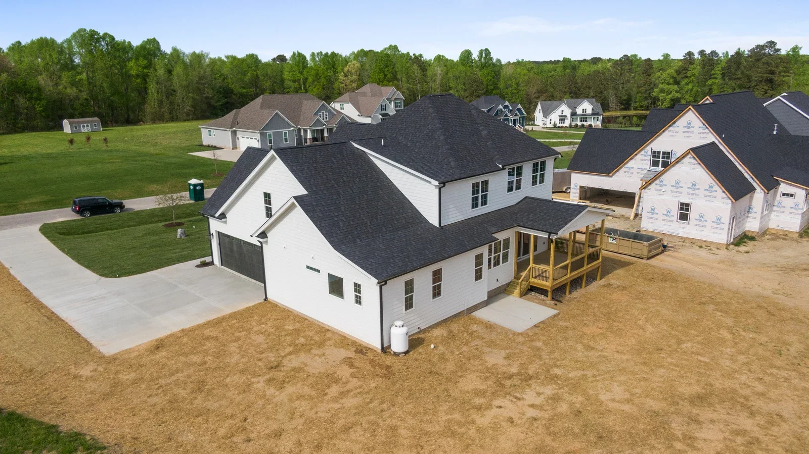 Aerial view of a newly built white two-story house with black roof shingles, a front porch, and a driveway, with other houses under construction and trees in the background.