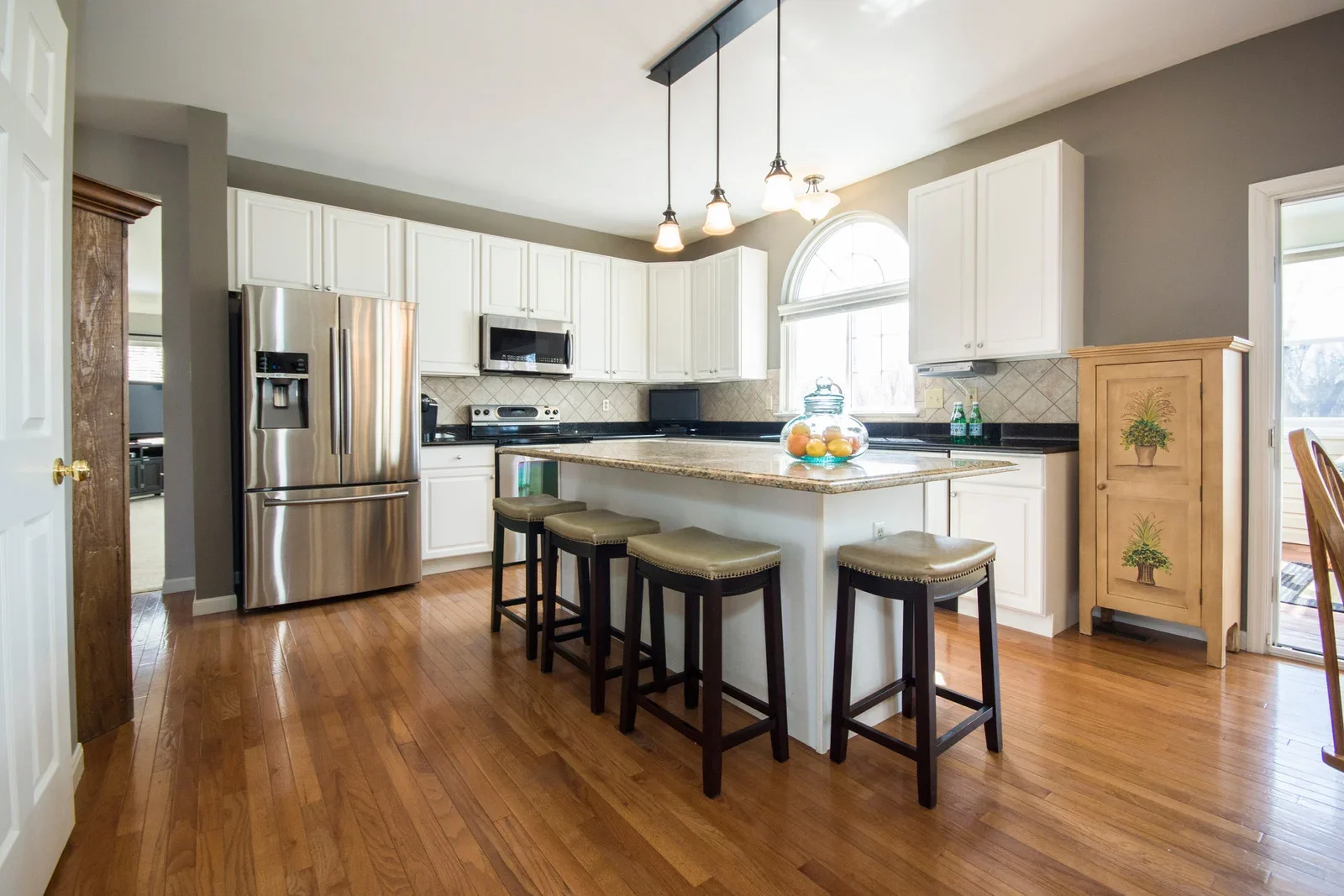 A kitchen with white cabinets, a stainless steel refrigerator, a black granite countertop, a microwave, and a breakfast bar with four stools. There is a window above the counter and a small wooden cabinet with painted potted plant designs.