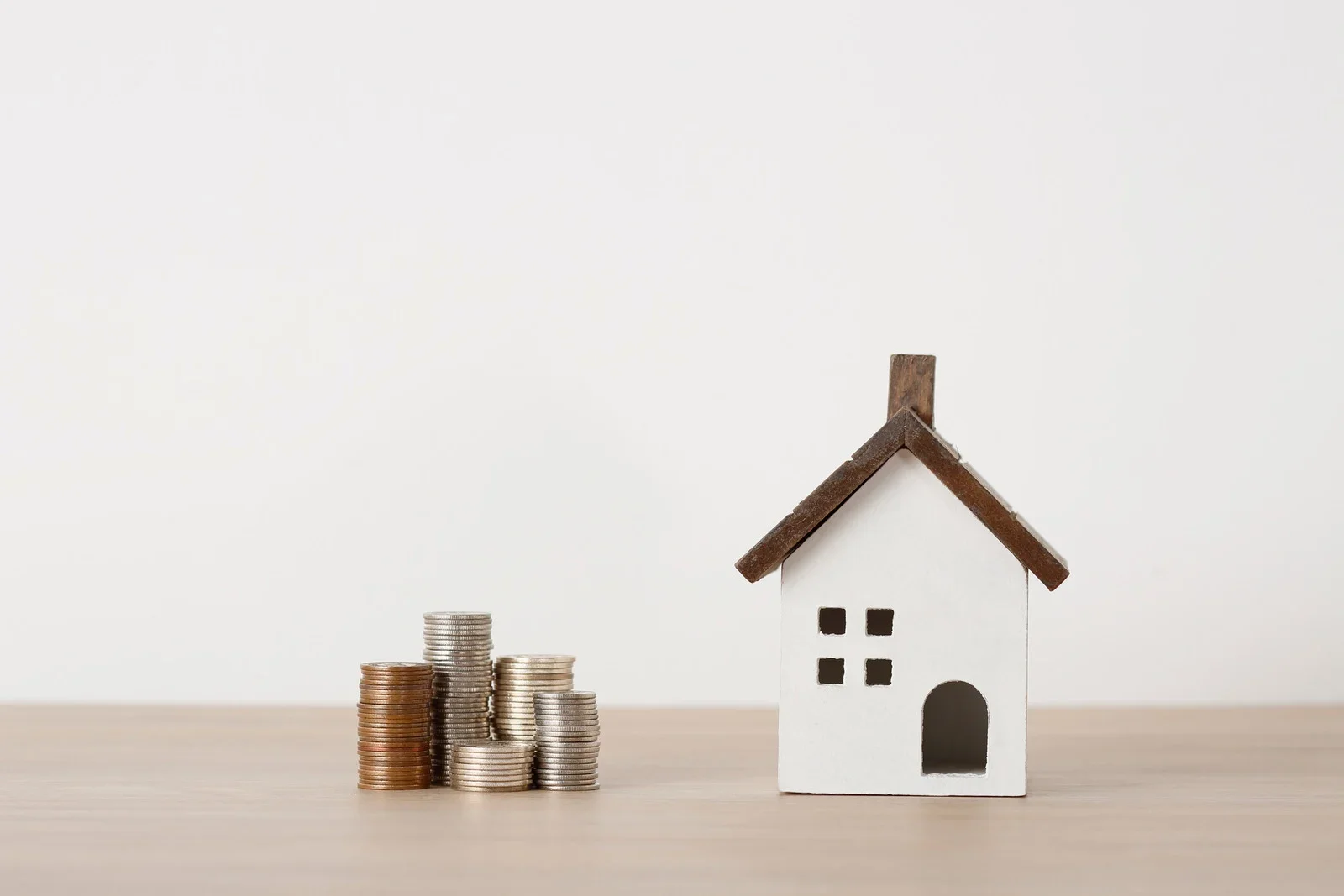 Stacks of coins next to a small white house model with a brown roof on a wooden surface.