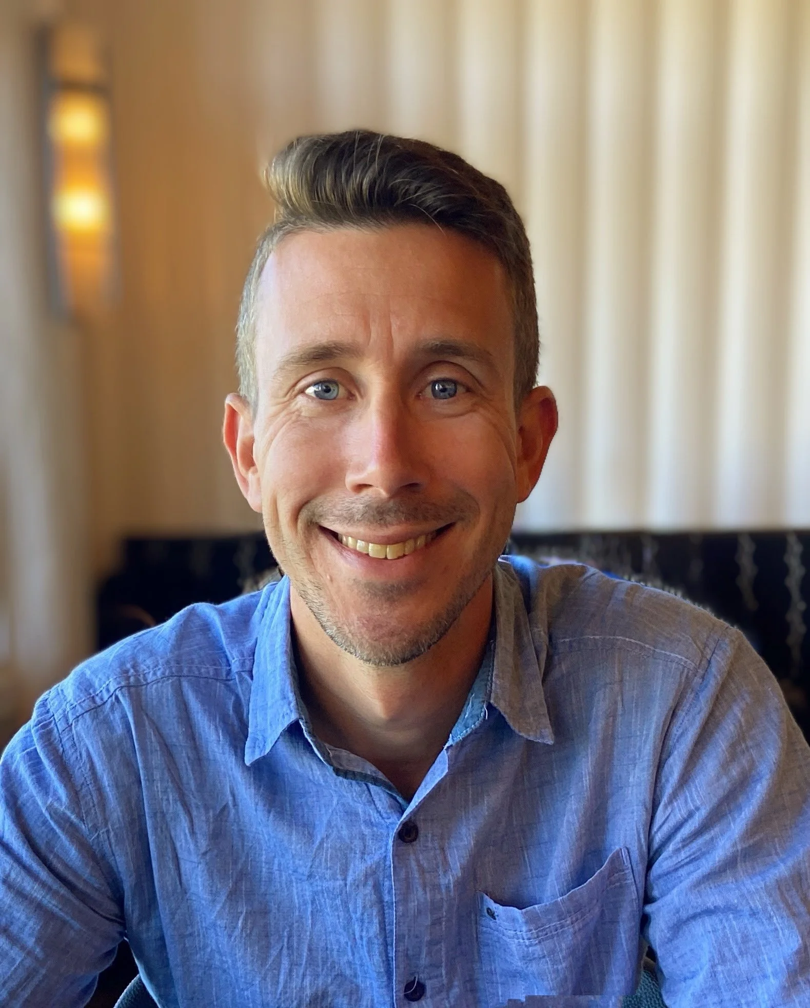 A smiling man with short brown hair and blue eyes, wearing a light blue button-up shirt, sitting in a well-lit room with cream-colored curtains and a dark piece of furniture in the background.