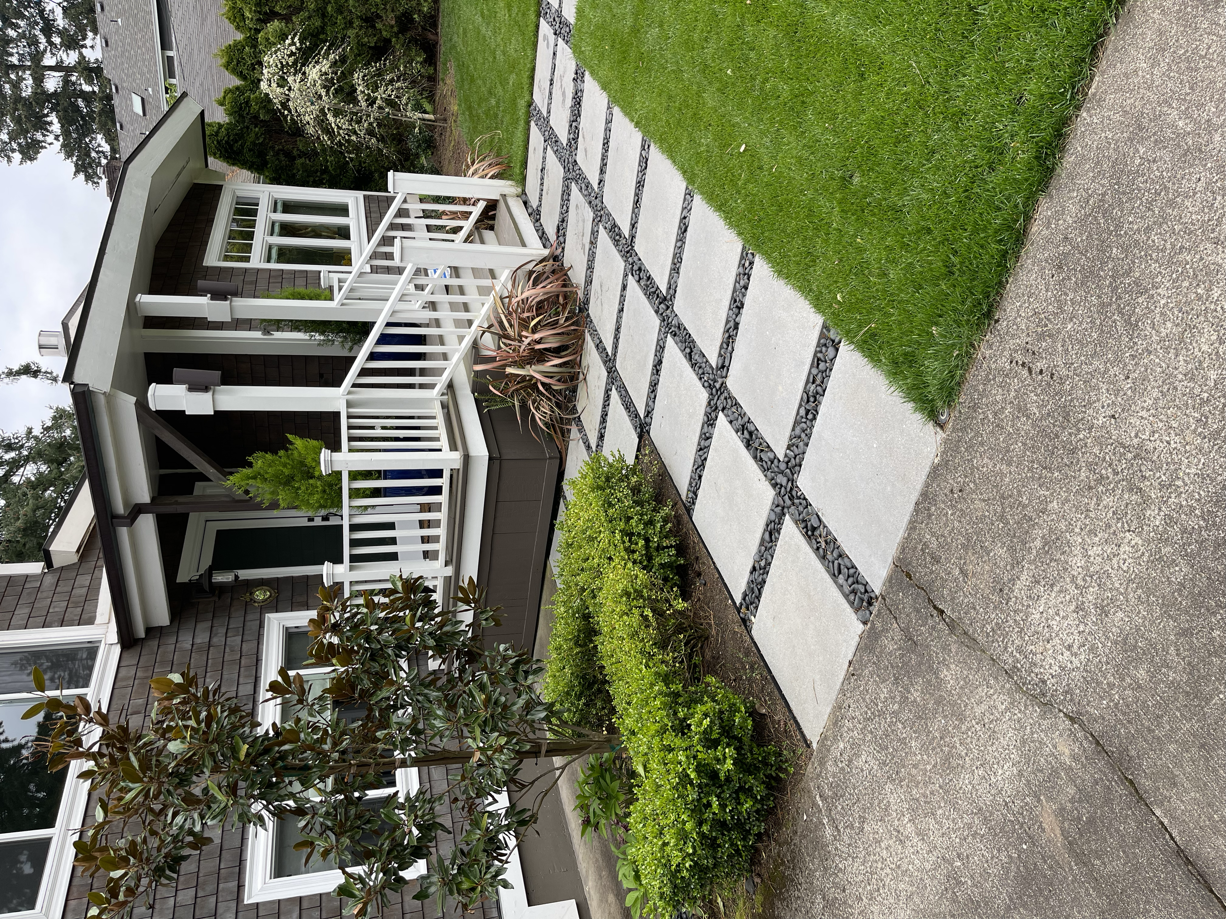 Front porch with white railing and steps, neatly paved walkway with square tiles and black stones, green plants and bushes, house with large windows, and a lawn.