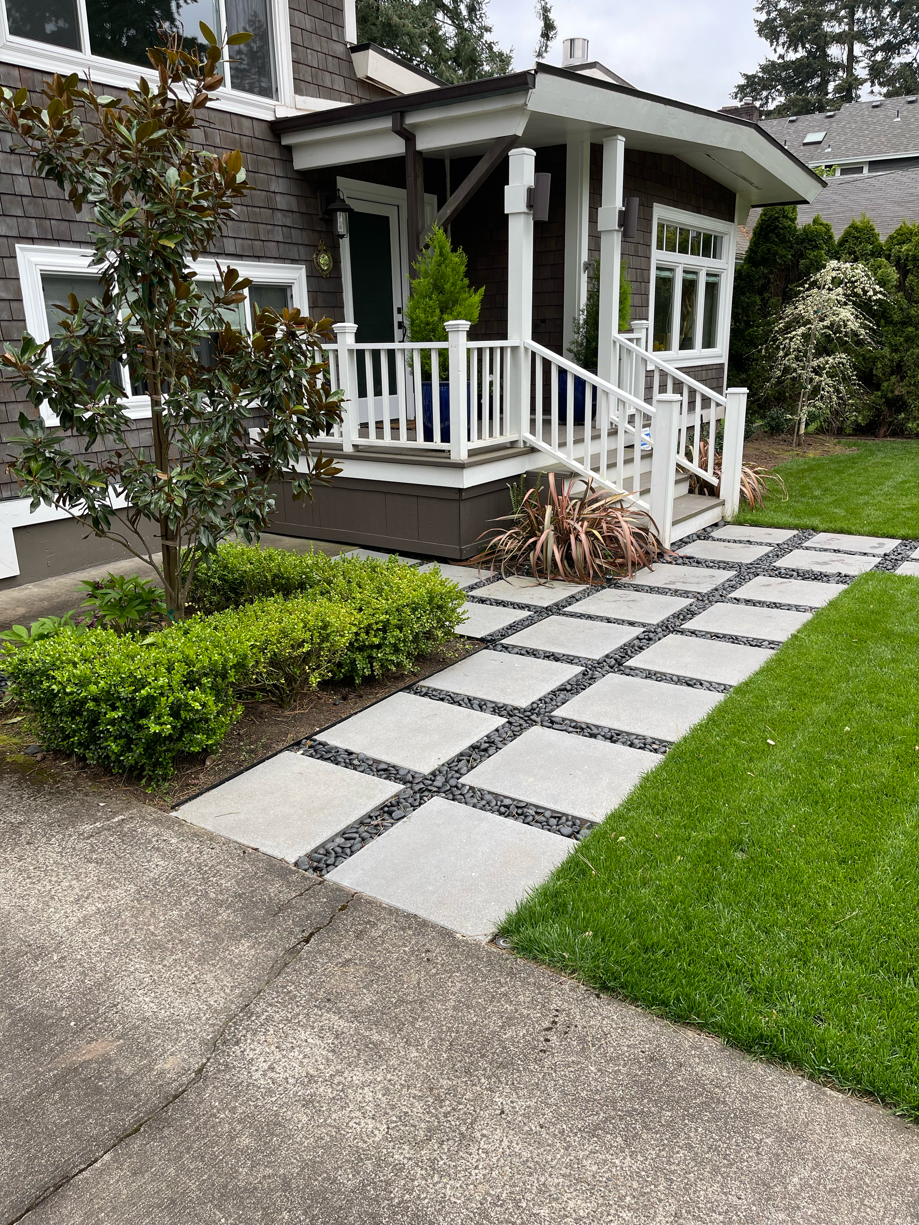 Front porch with white railing and steps, neatly paved walkway with square tiles and black stones, green plants and bushes, house with large windows, and a lawn.