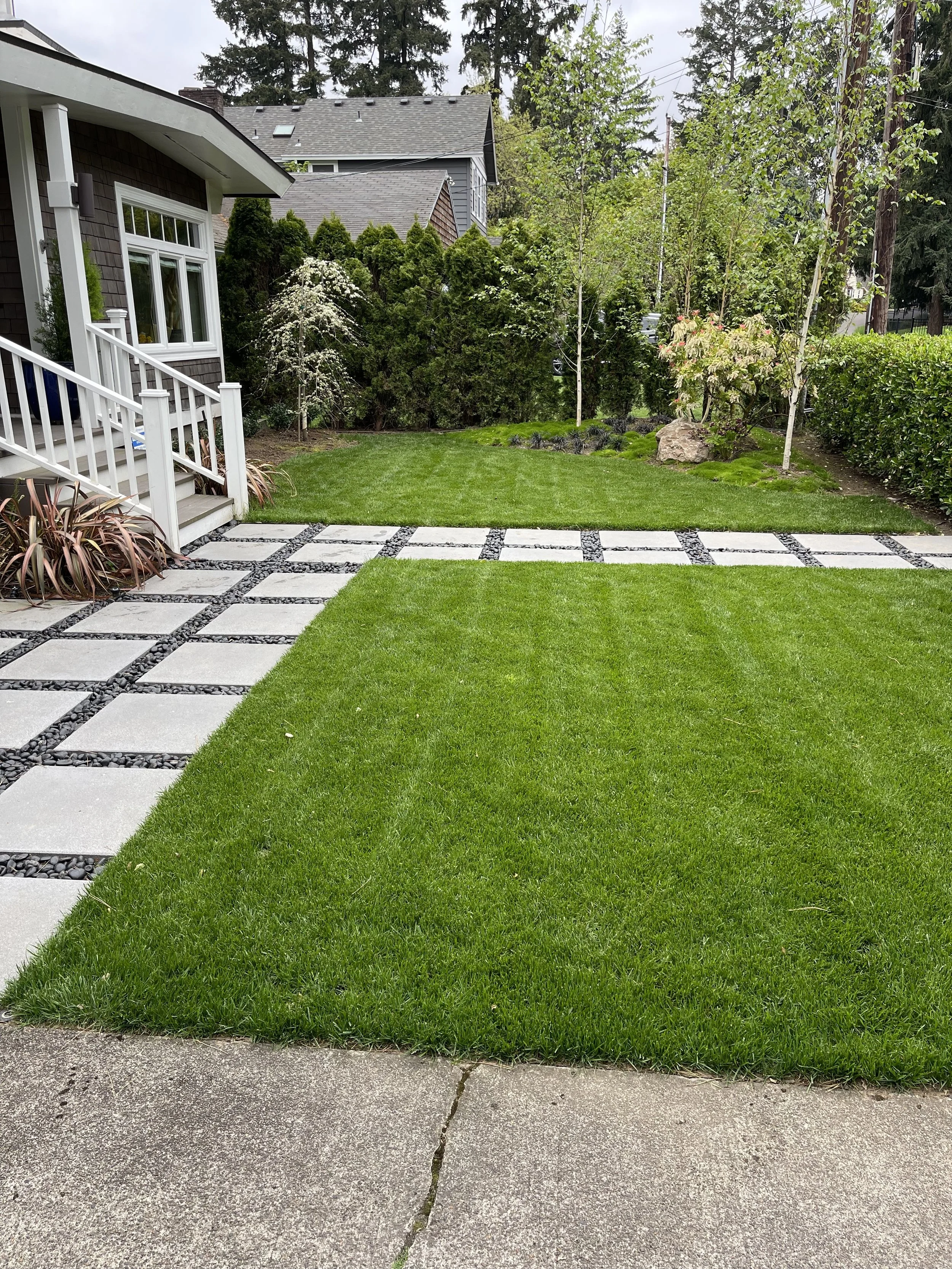 A backyard with a well-maintained lawn, concrete stepping stones with black gravel in between, a white staircase leading to a porch, and various trees and bushes along the fence line.
