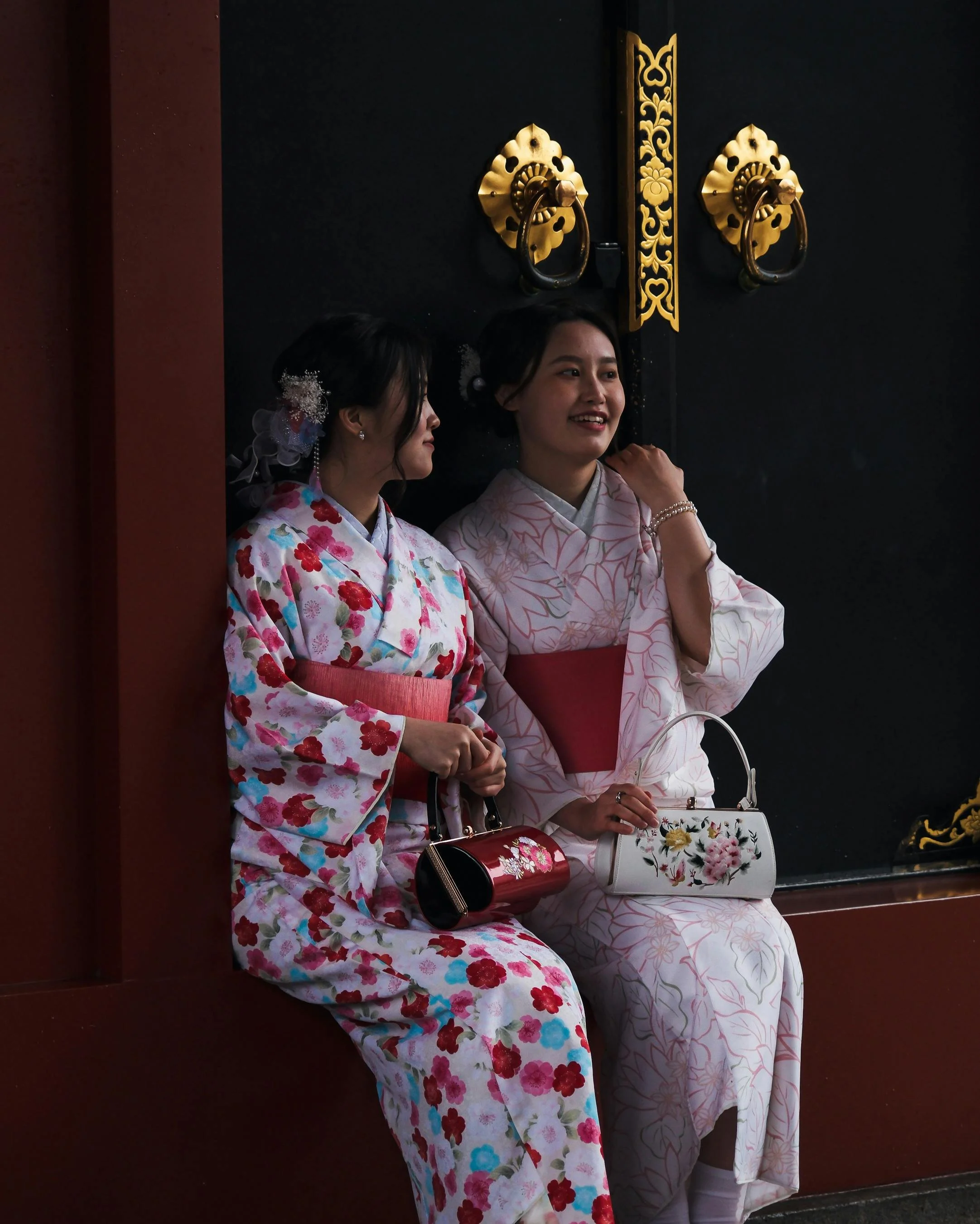 Two women in traditional Japanese kimonos sitting against a black door with ornate gold door handles and decorative gold accents.