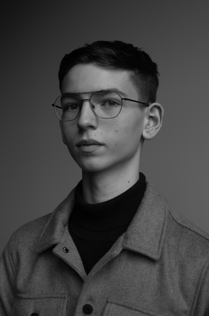 Black and white portrait of a young man with short hair, wearing glasses, a dark turtleneck, and a light jacket, looking directly at the camera.
