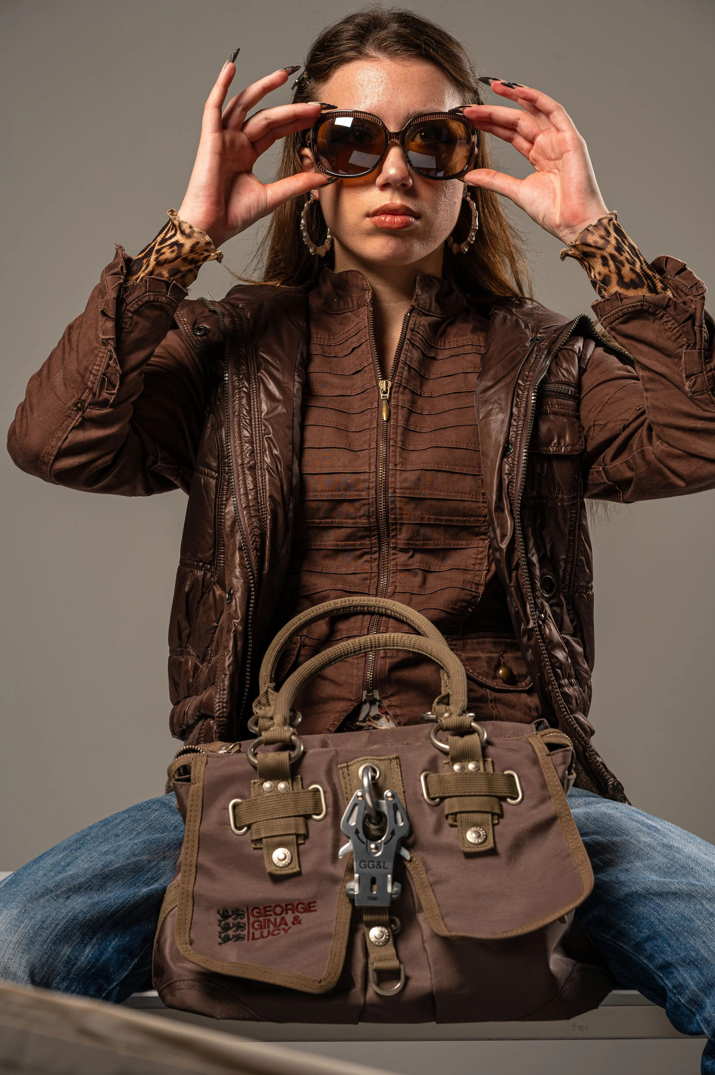 A woman wearing sunglasses, a brown leather jacket, and a ruffled brown shirt. She is sitting with legs apart and holding her sunglasses with both hands, with a beige bag featuring red and black embroidery and metal hardware in front of her.