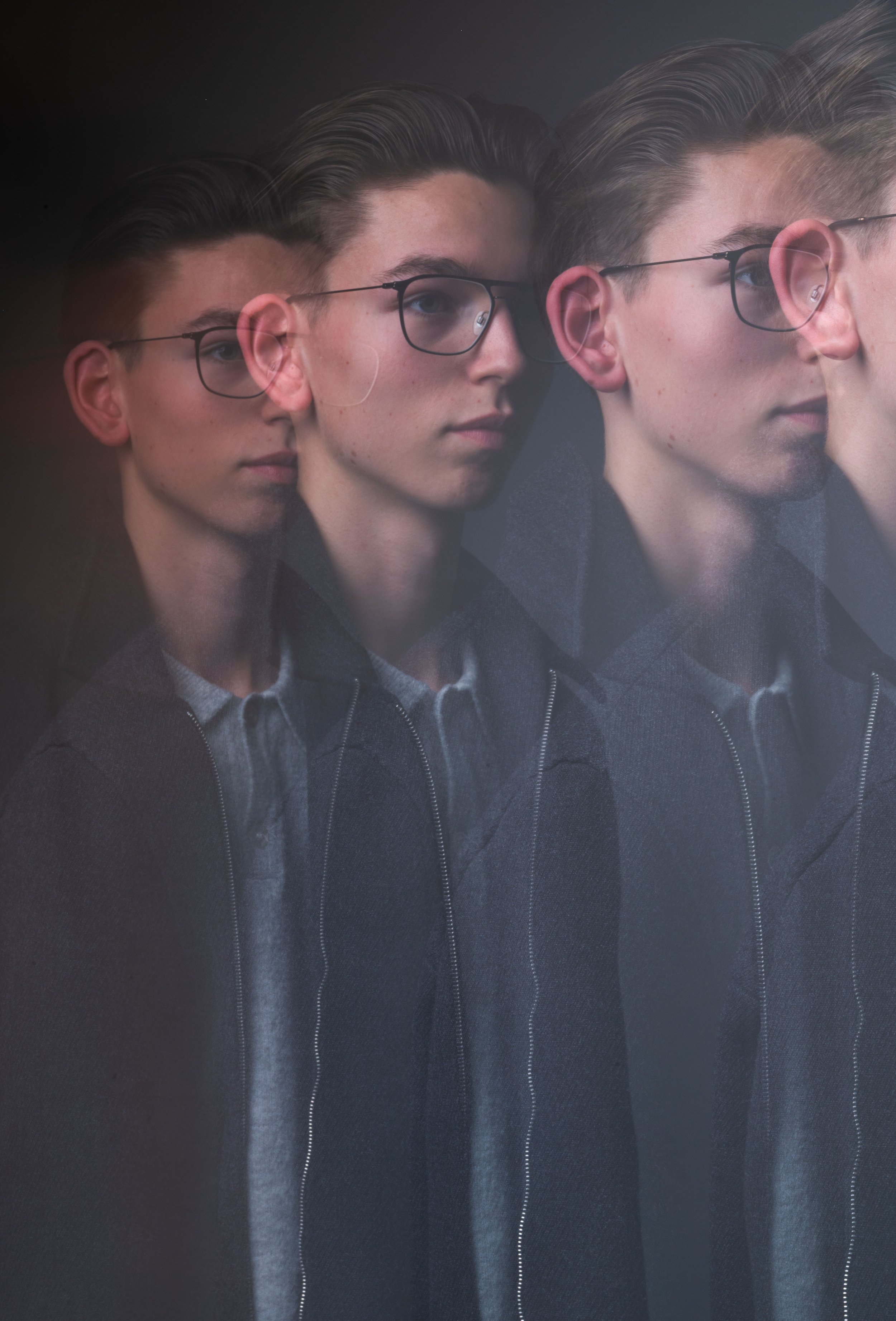 Multiple overlapping images of a young man with blond hair, glasses, wearing a dark jacket and collared shirt, creating a glitch-like effect.