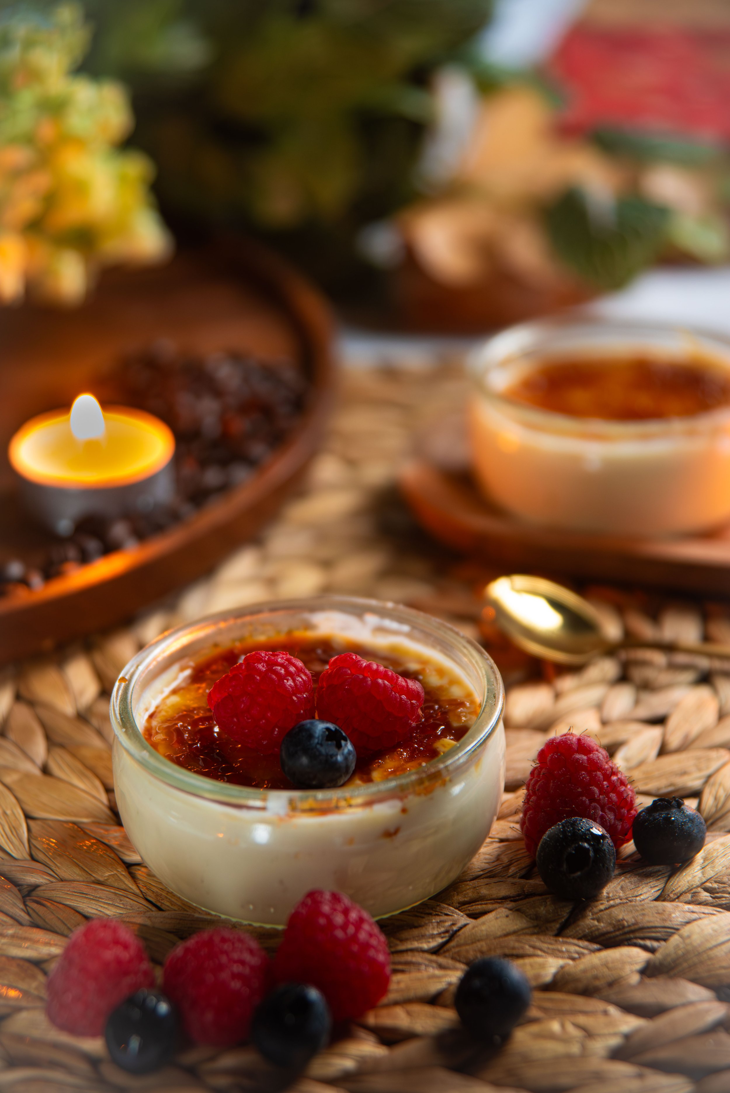 Dessert cups with berries on a woven tray, with a lit candle and flowers in the background.