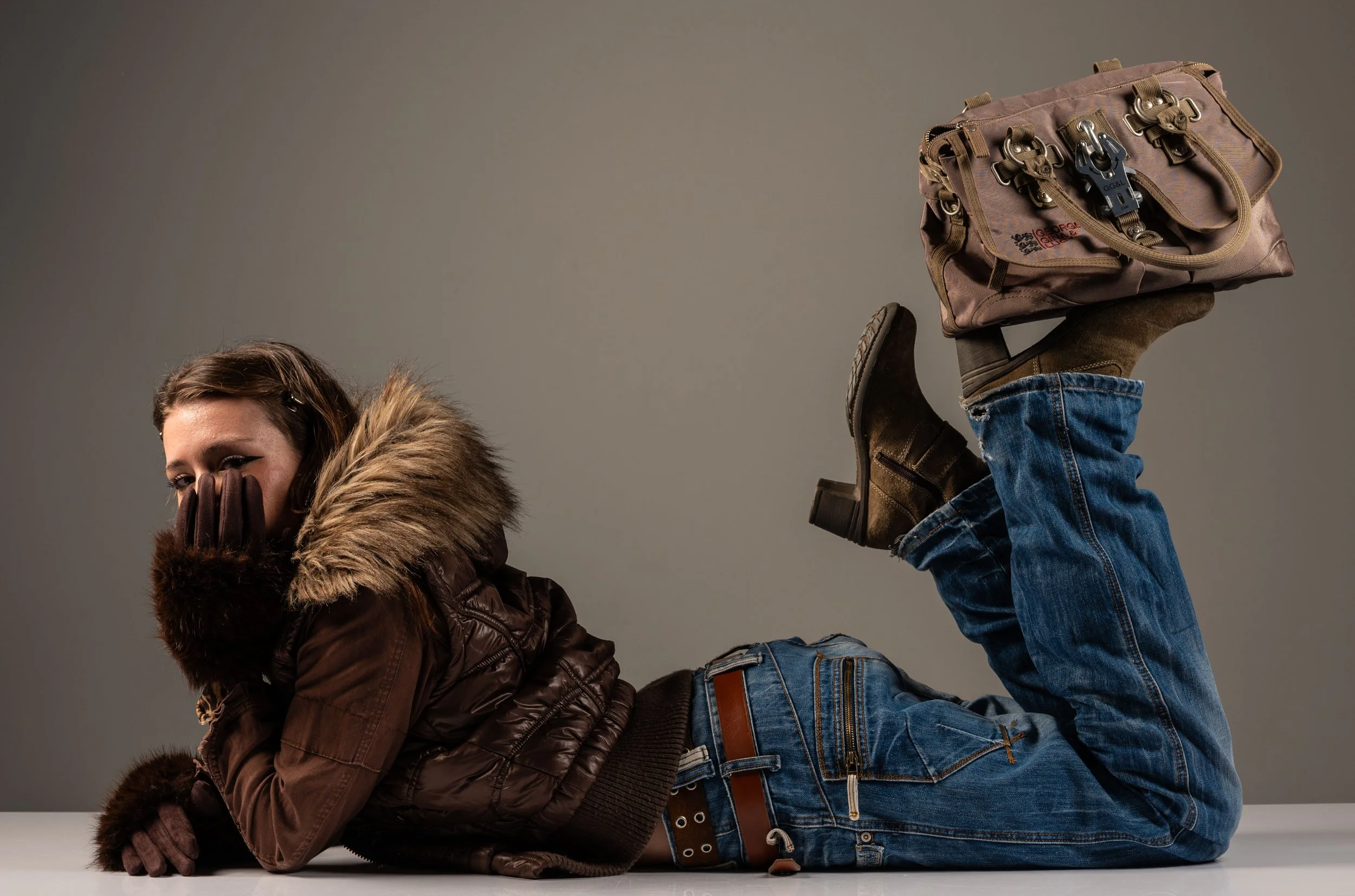 A young woman is lying on her stomach with her legs raised, wearing a brown winter jacket with fur trim, blue jeans, and brown boots, holding a beige bag with multiple locks and zippers while covering her face with her hand.