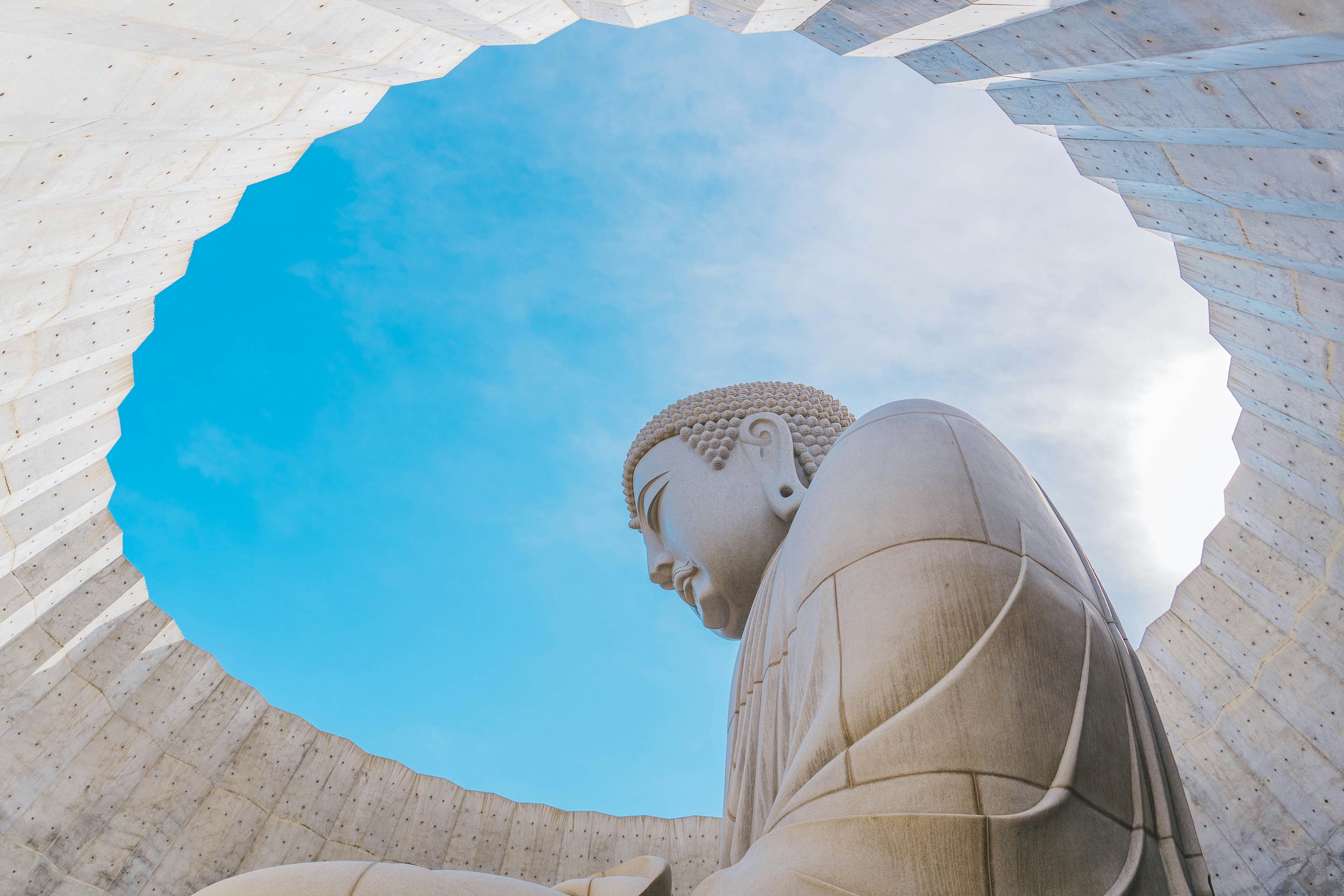 The image shows the large carved face of a seated Buddha statue viewed from below, framed by an open circular stone structure with a blue sky in the background.