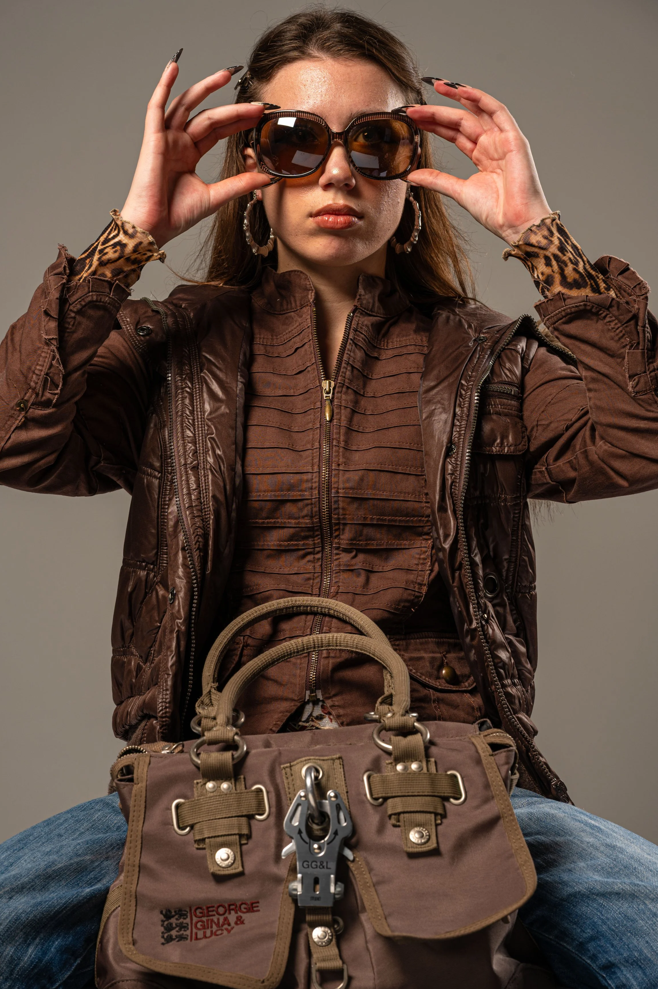 A young woman with long brown hair wearing large sunglasses, hoop earrings, a brown jacket with a zip, a patterned top, and blue jeans, sitting with a neutral background, holding her sunglasses with both hands, and a tan bag with patches and hardware resting on her lap.