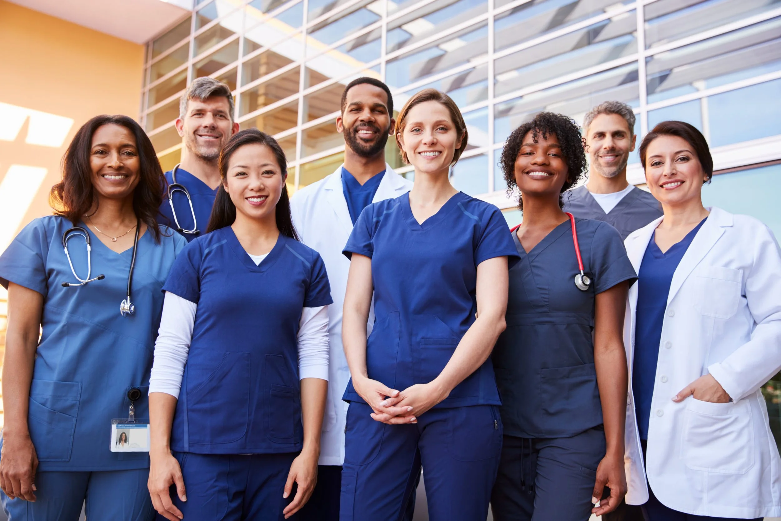 A diverse group of nine healthcare professionals in scrubs and lab coats, standing outside a medical facility, smiling at the camera.