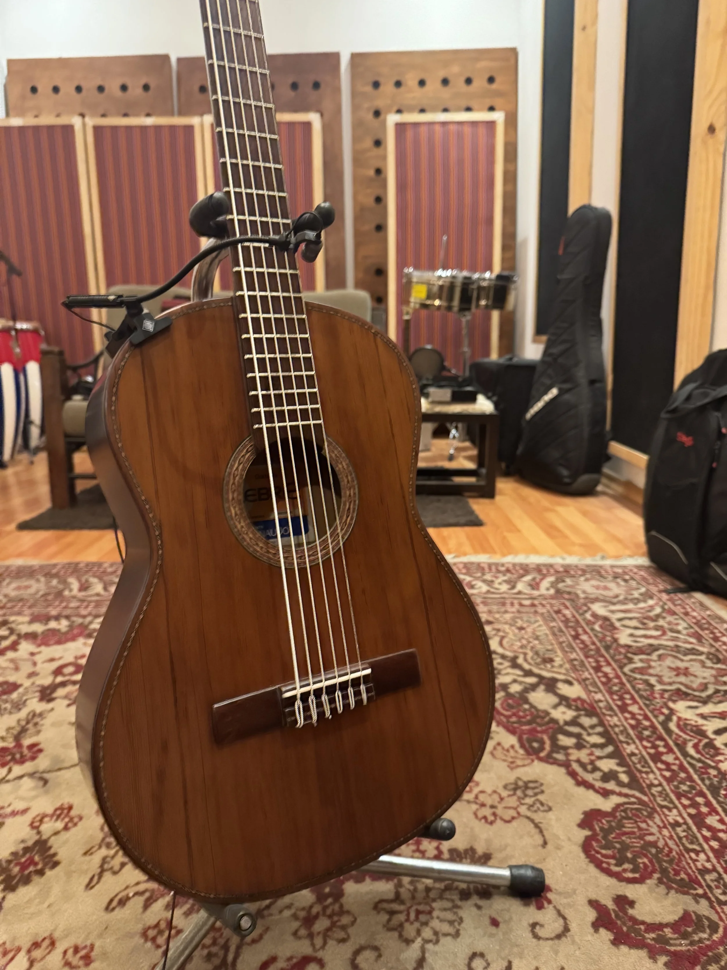 An acoustic guitar on a stand in a recording studio with soundproof panels, musical instruments, and equipment in the background.