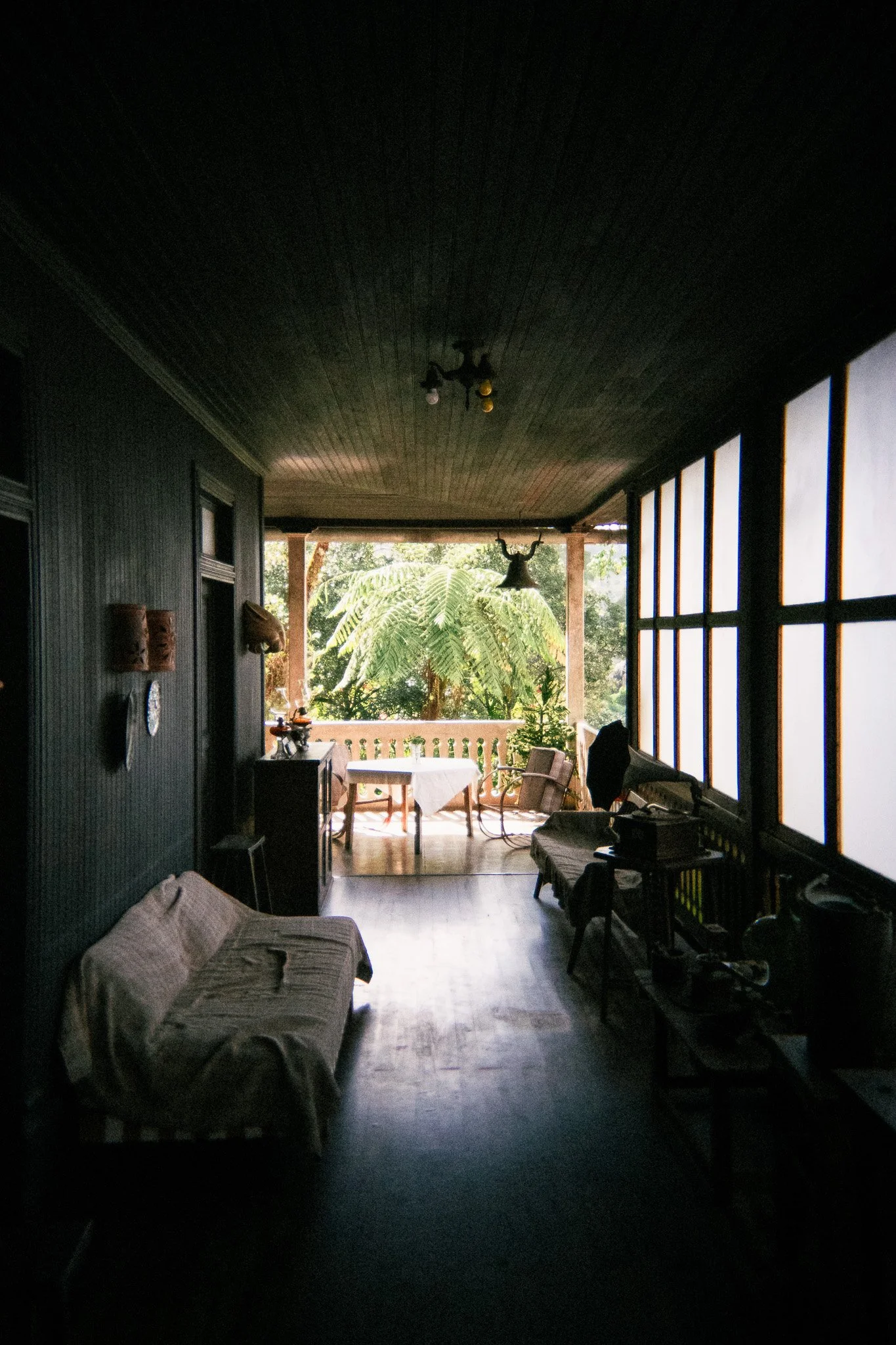 Interior of a dark room with a wooden ceiling, leading to a bright balcony with trees outside. There is a sofa with a blanket, and a table with chairs on the balcony.