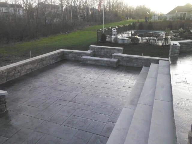 Empty backyard patio with gray stone flooring, steps leading down, and a low stone wall with a seating area, fence, and grassy yard in the background.
