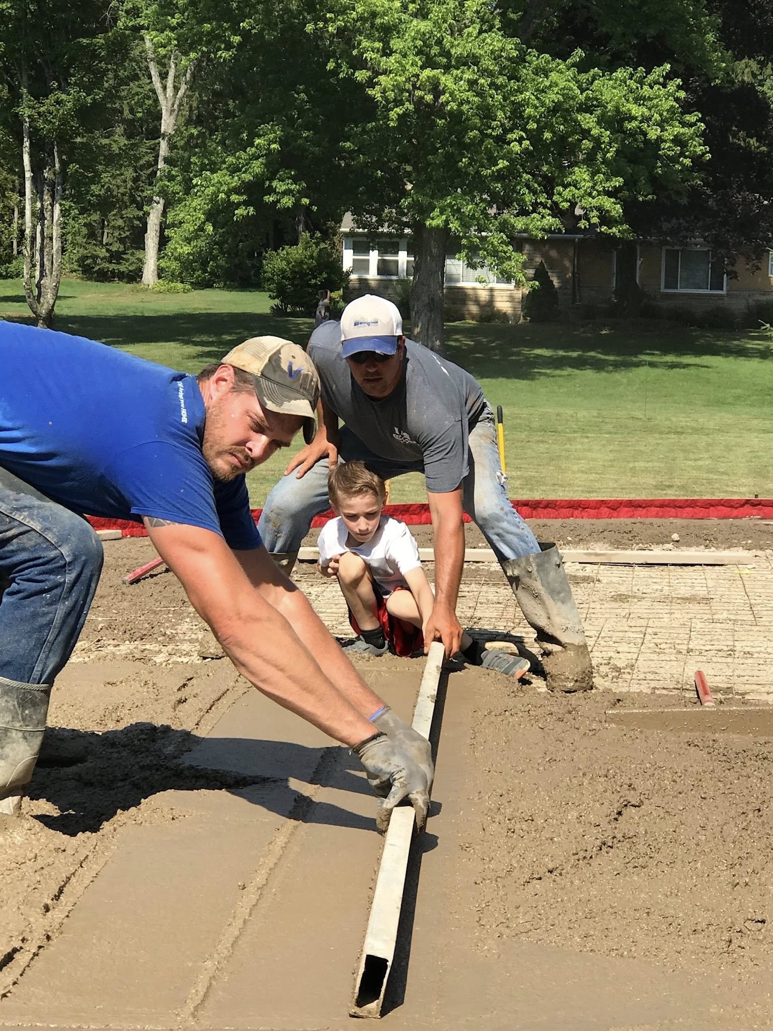 Three people, two men and a child, are working together on a construction site to level wet concrete with a long straightedge. The setting is outdoors with green trees and a building in the background.