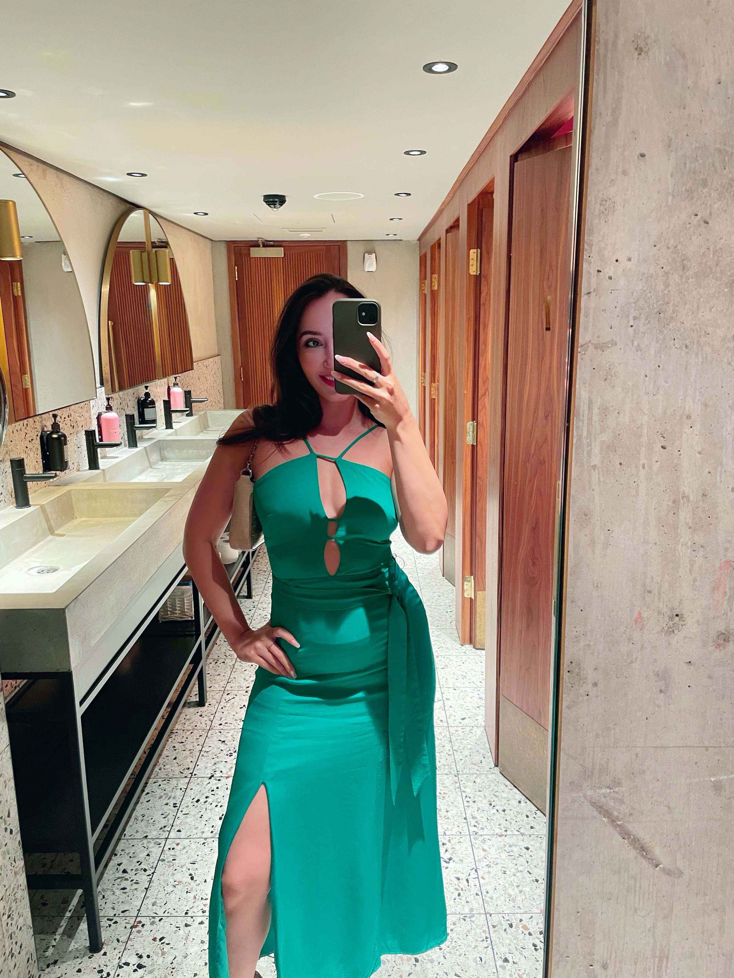 A woman in a teal dress taking a mirror selfie in a bathroom with wood-paneled doors and modern sinks.