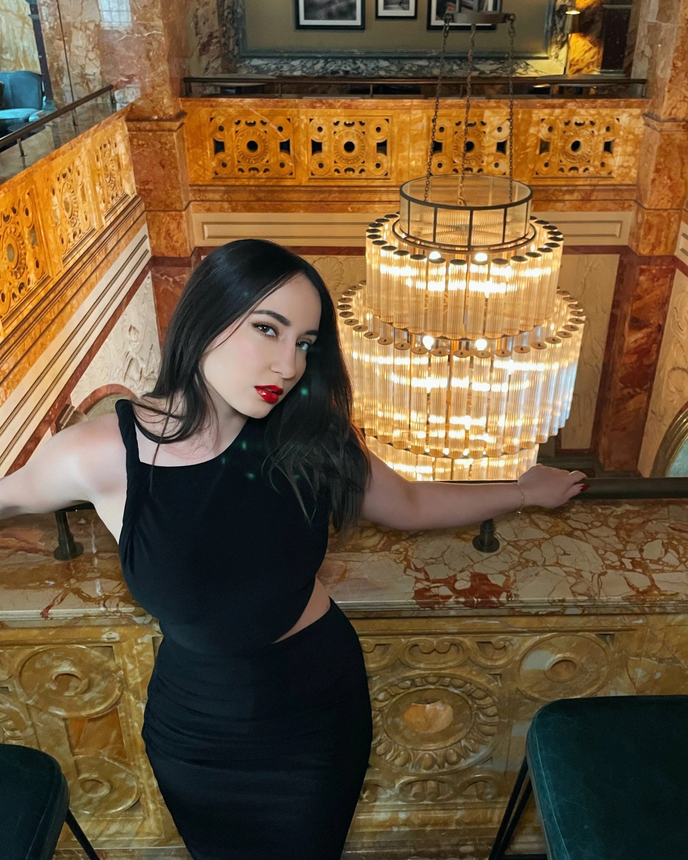 Miss Kat is an Elite Companion in London  with long dark hair and red lipstick posing on a staircase railing inside an ornate, luxurious hotel or mansion with marble walls and golden decorative details, and a large glowing chandelier.