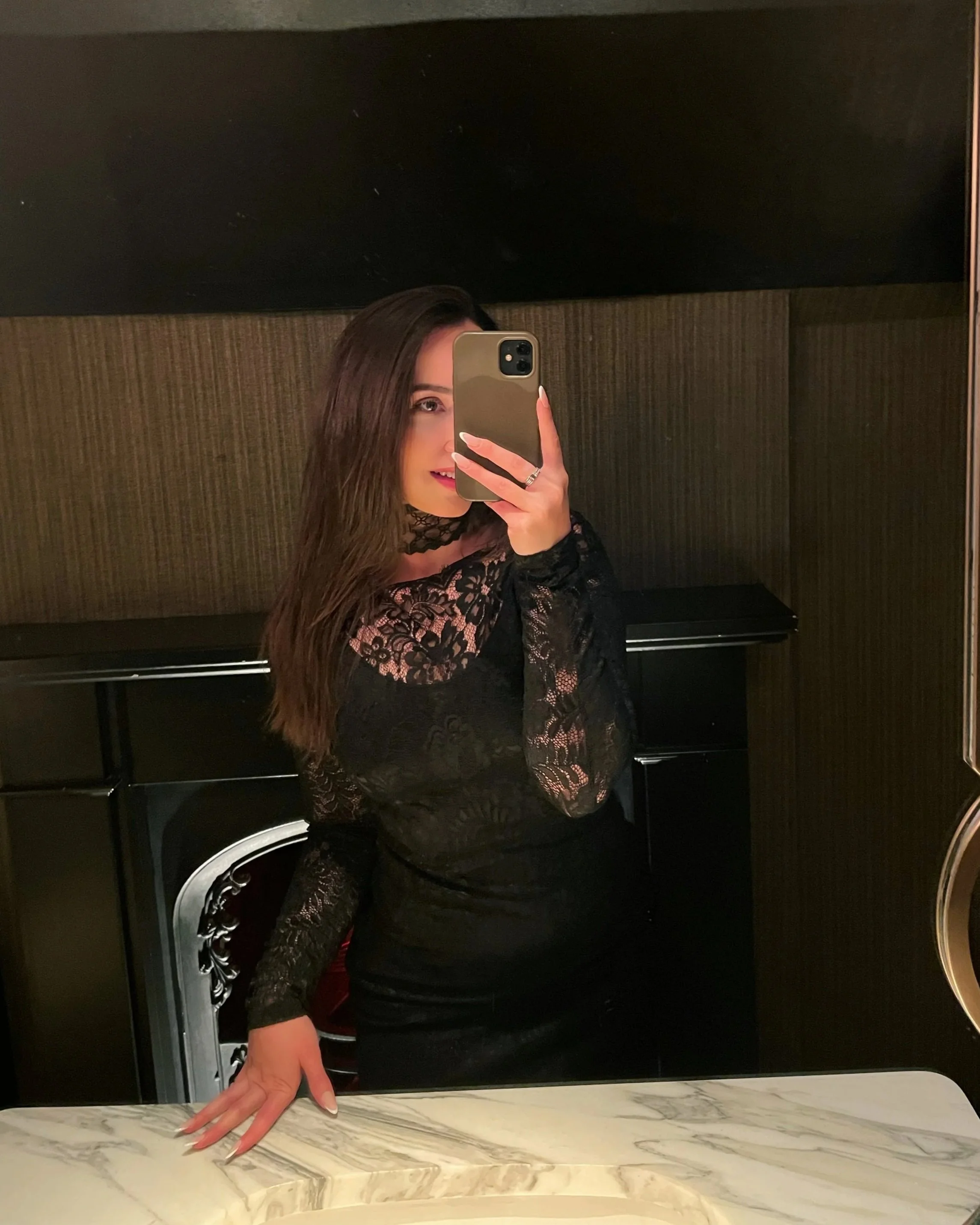 A woman with long dark hair wearing a black lace dress taking a mirror selfie with a smartphone in a room with dark walls and a marble countertop.