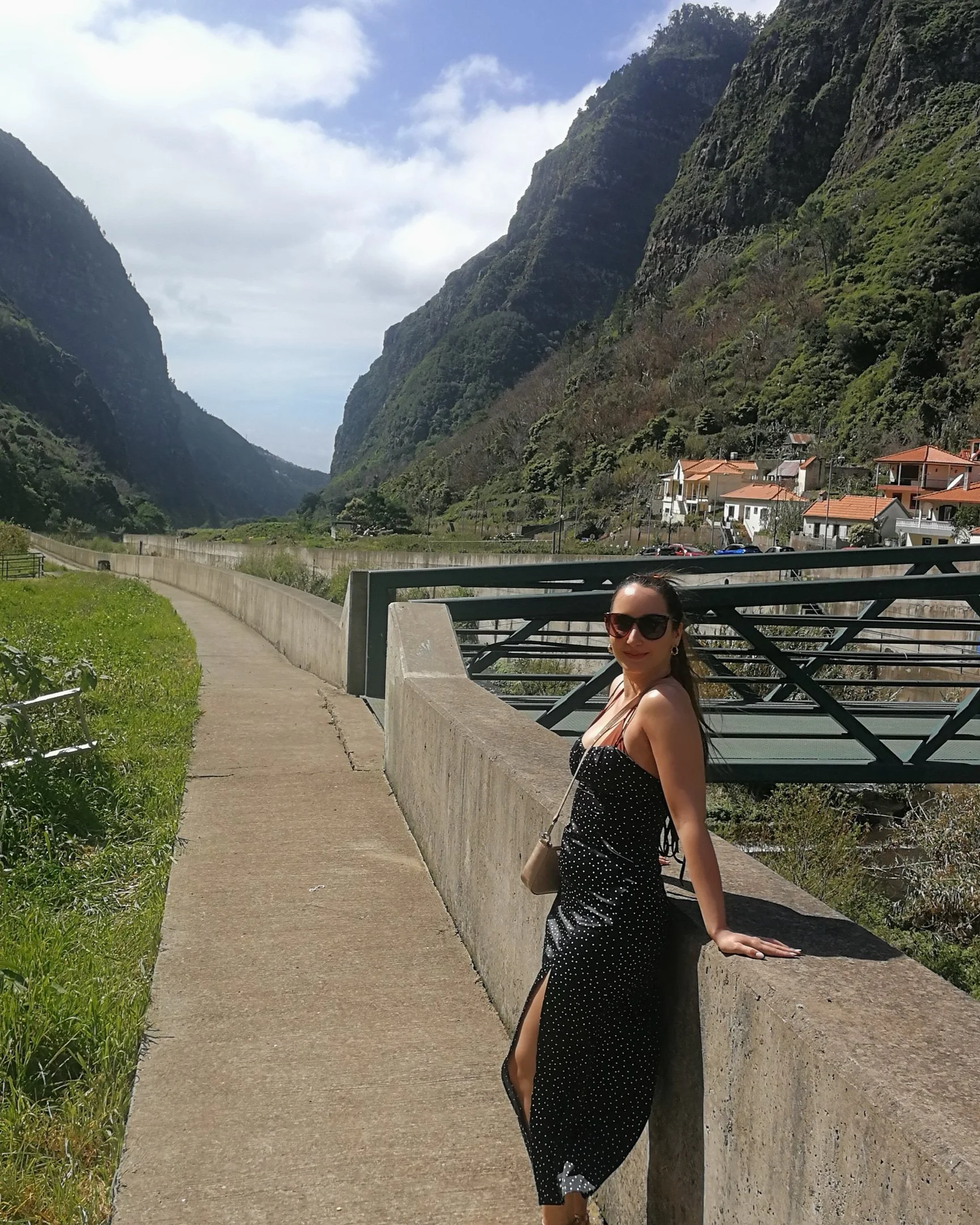 A woman in a black polka dot dress and sunglasses leaning on a roadside wall with mountains and houses in the background on a partly cloudy day.