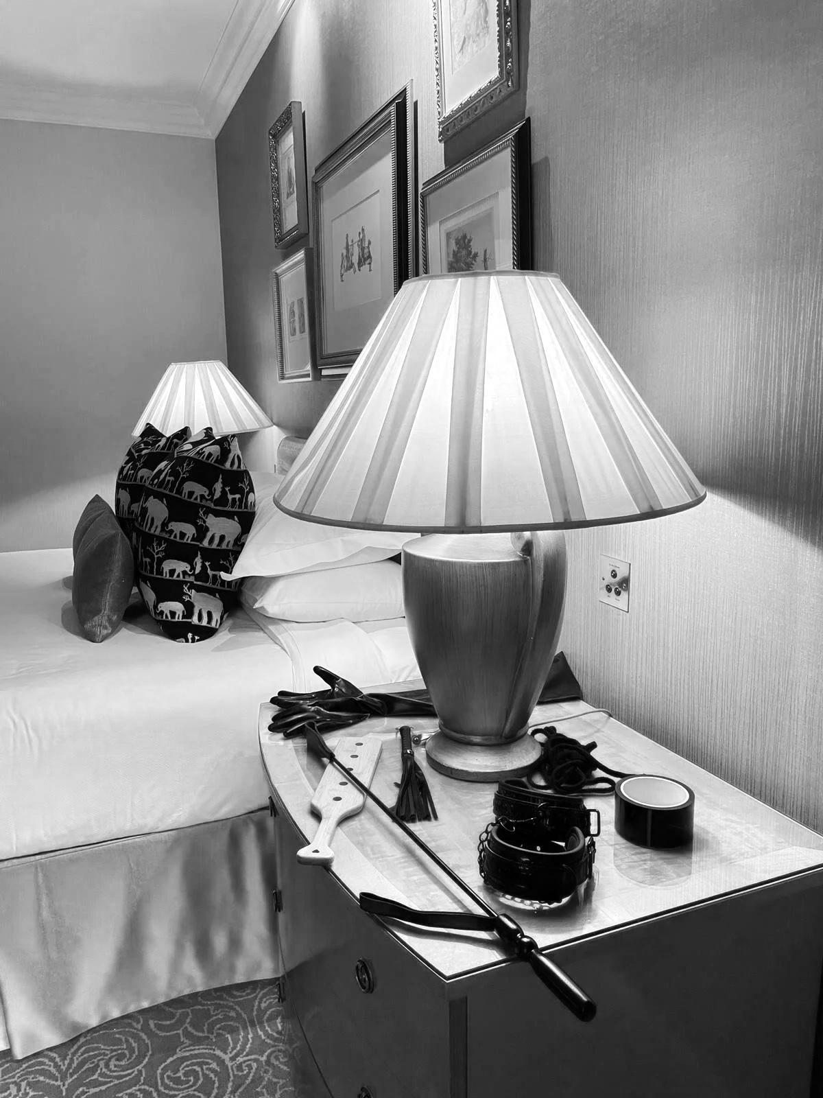 A bedside table with a large table lamp, gloves, belt, and other items. A bed with patterned pillows and framed pictures on the wall above.